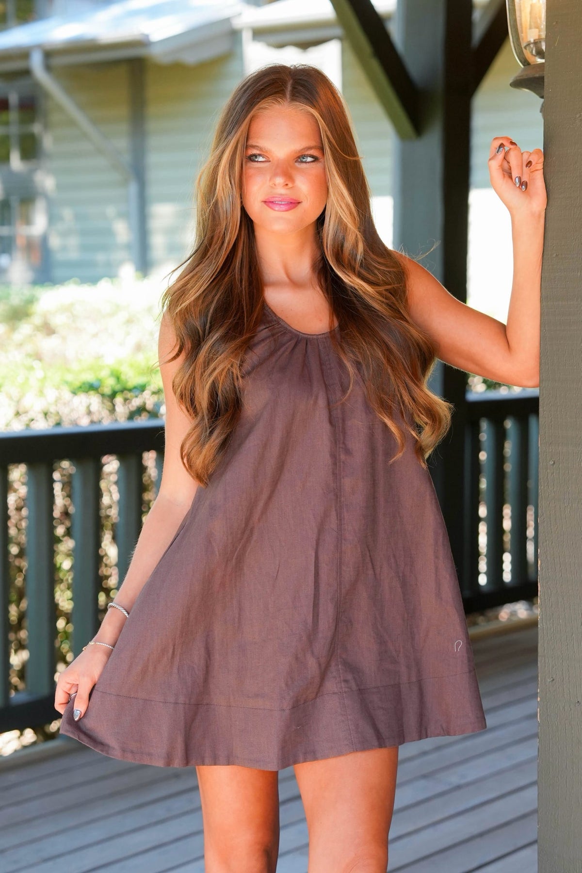 A woman wearing a sleeveless, scoop-neck, A-line mini dress in a dusty mauve-brown color, standing on a wooden deck.