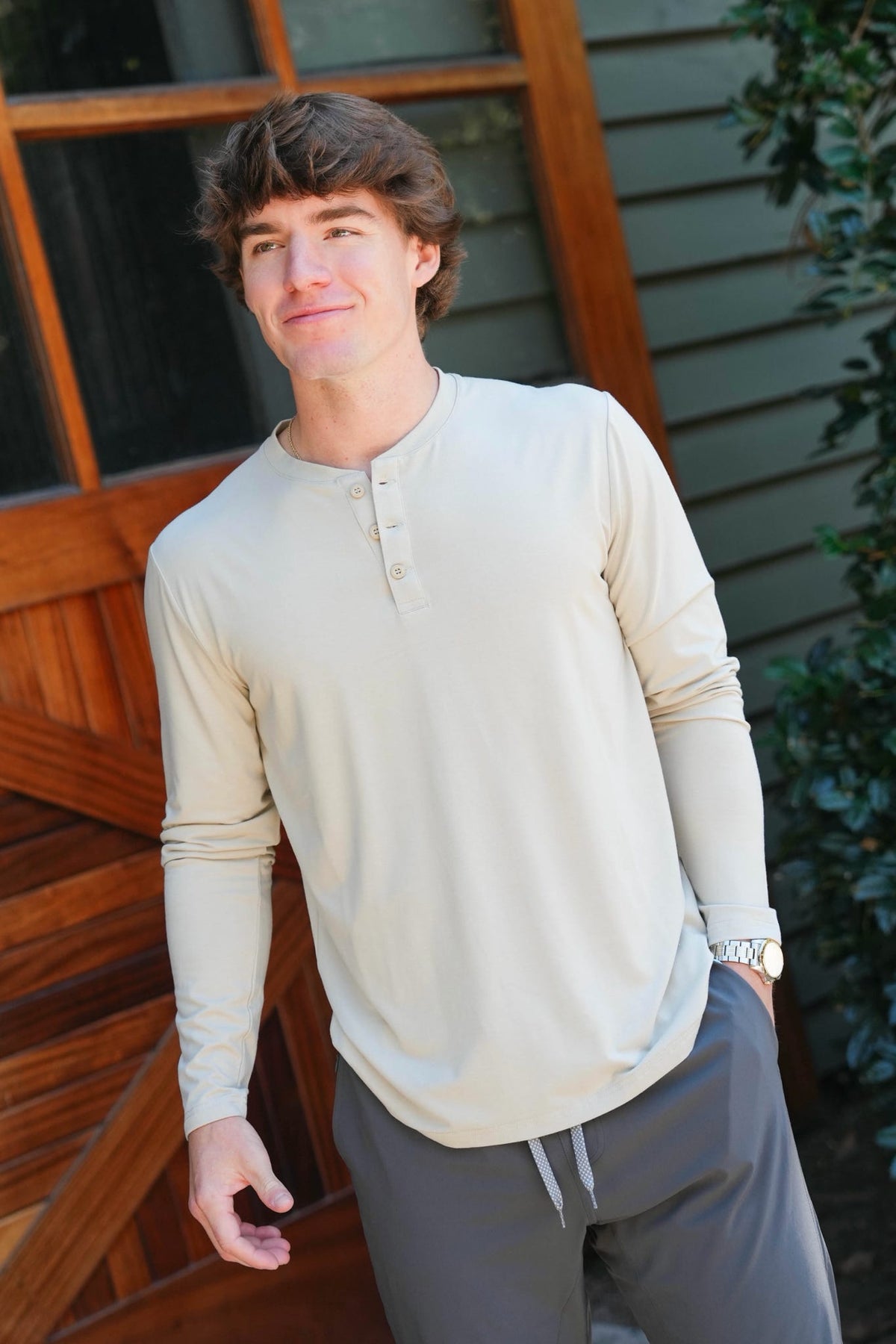 A man wearing a light beige long-sleeve henley shirt and gray drawstring pants, standing outdoors in front of a wooden door and green foliage.