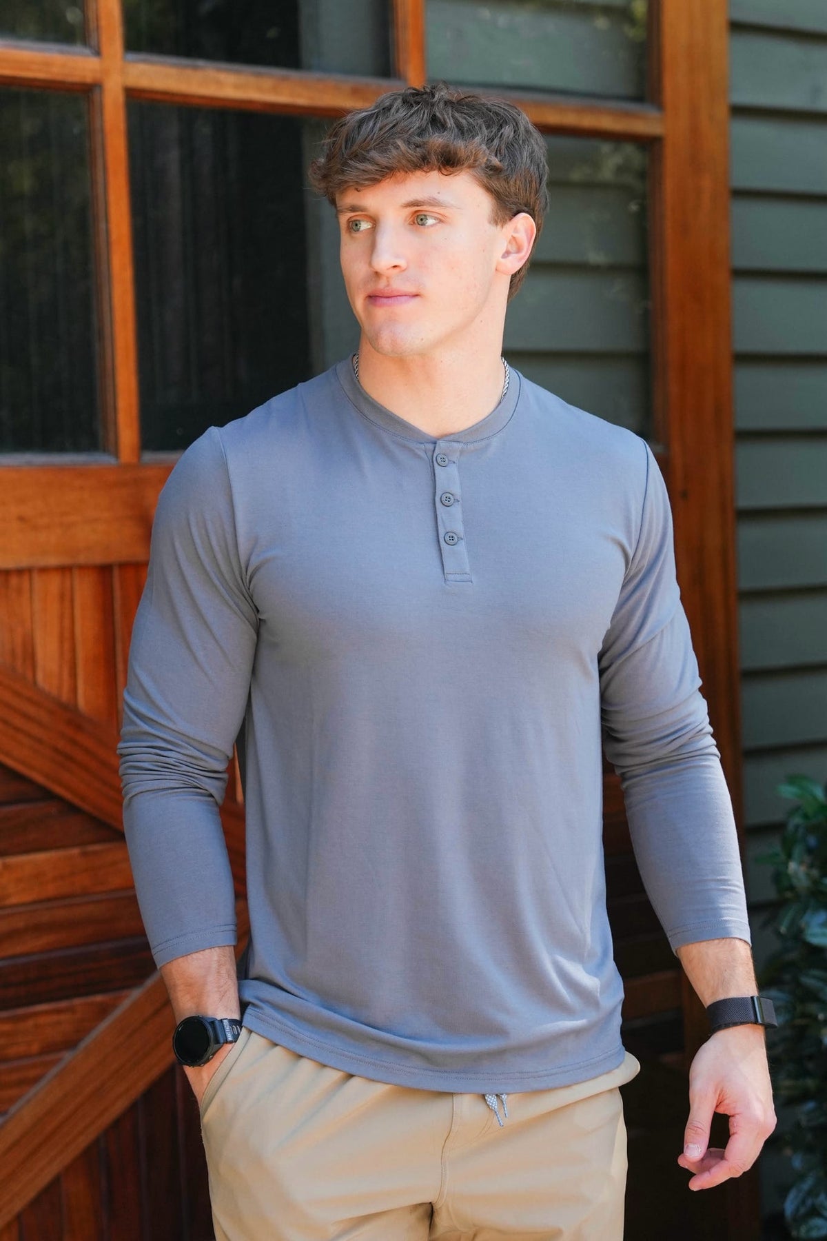 A man wearing a gray long-sleeve henley shirt and khaki pants, standing in front of a wooden door and house siding.
