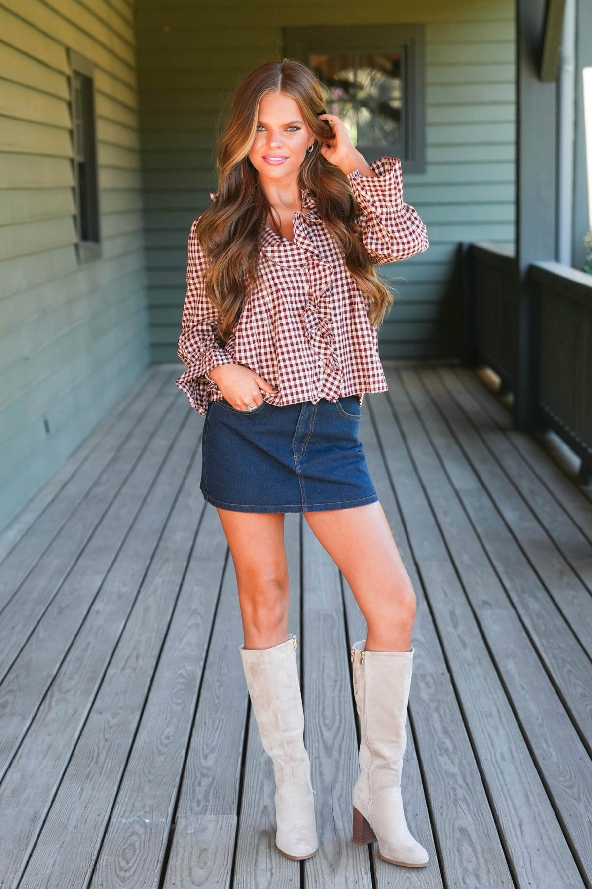 A woman stands on a wooden porch wearing a red and white gingham long-sleeve blouse, a dark blue denim mini skirt, and off-white knee-high boots.
