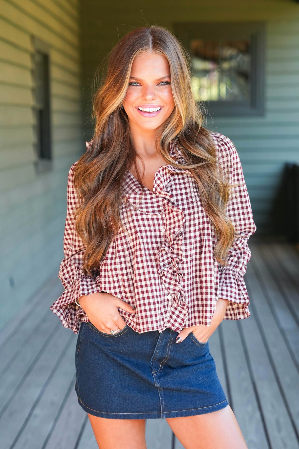 A woman with long wavy brown hair stands on a wooden porch wearing a red and white gingham ruffled blouse and a dark blue denim mini skirt.