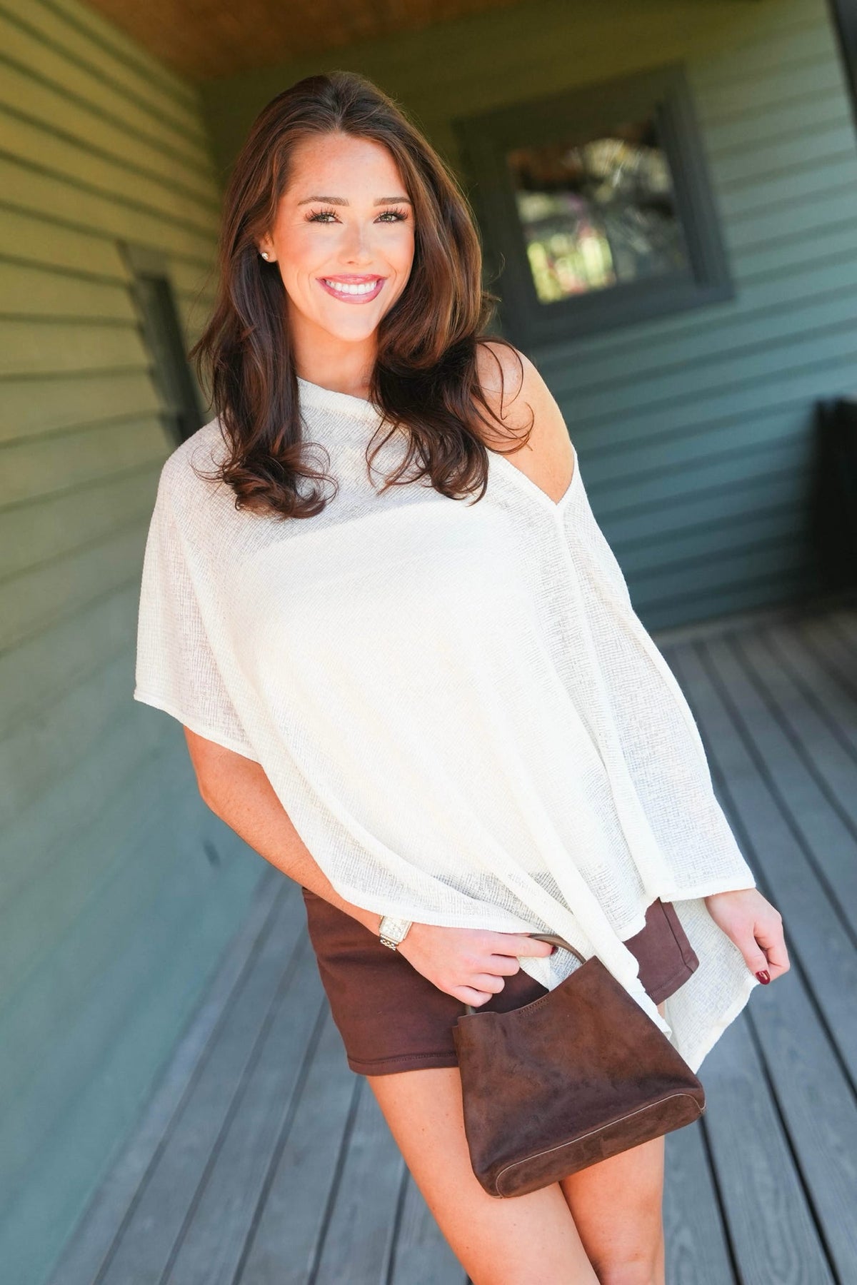 A woman wearing a white off-the-shoulder knit top and brown shorts, holding a brown suede clutch bag, standing on a wooden deck in front of a house.