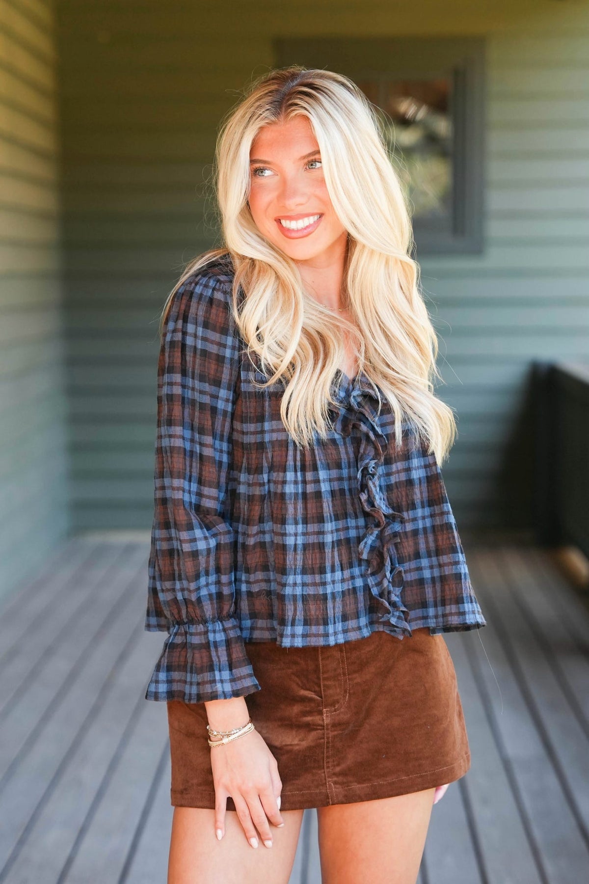 A smiling blonde woman wearing a brown and blue plaid long-sleeve blouse with ruffle details and a brown mini skirt, standing on a wooden deck in front of a house.