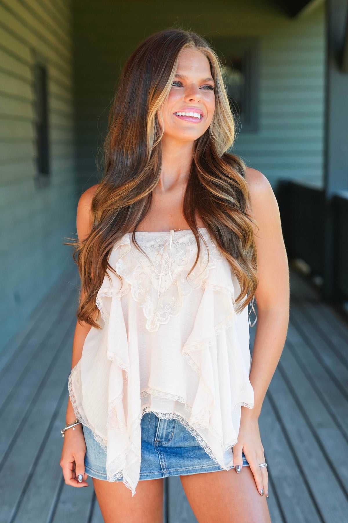 A woman with long brown hair models a sleeveless, ruffled cream top and denim shorts on a wooden porch.
