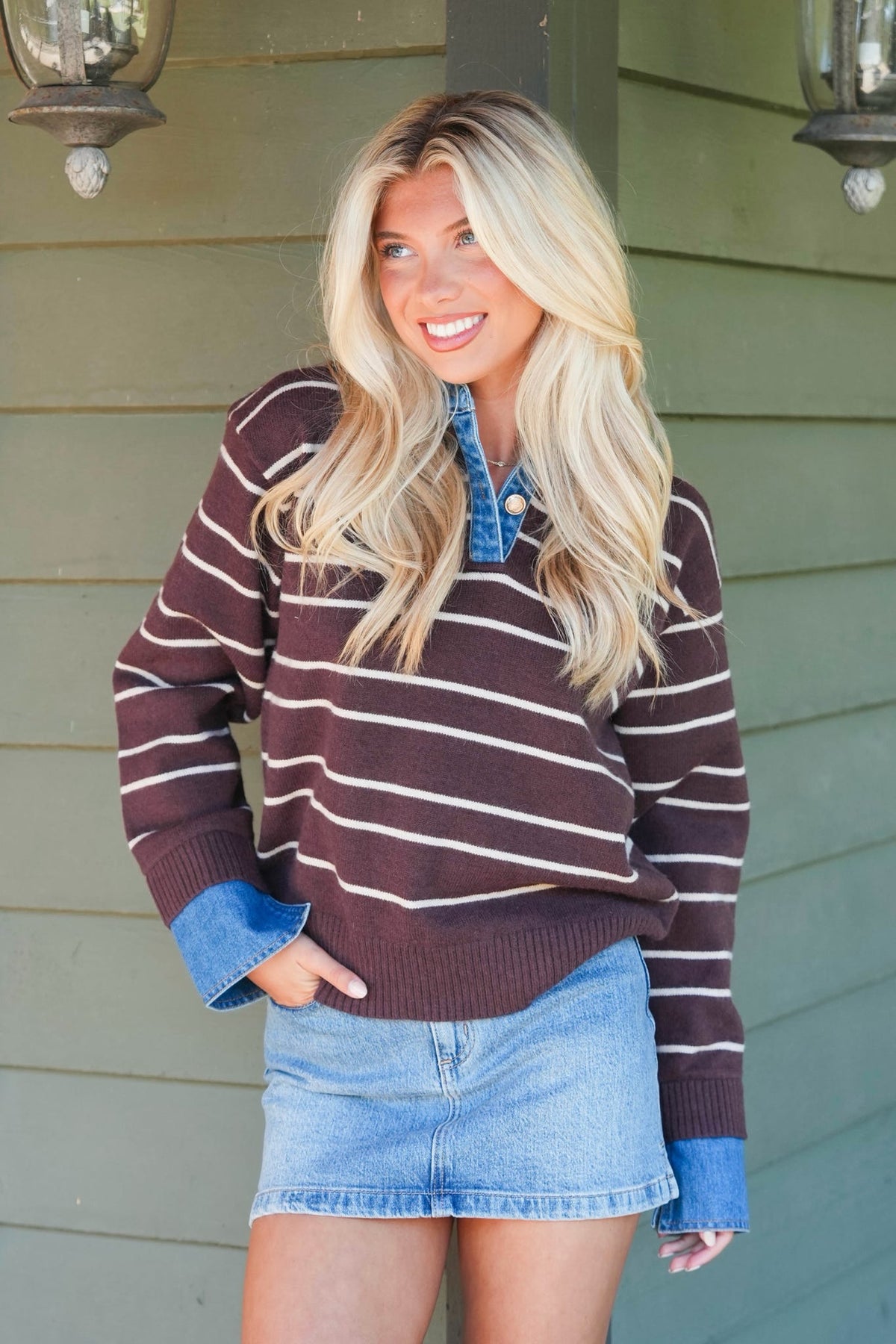 A smiling blonde woman wearing a brown and white horizontally striped knit sweater with blue button-up collar and cuffs, paired with a denim mini skirt, standing in front of a green house exterior.