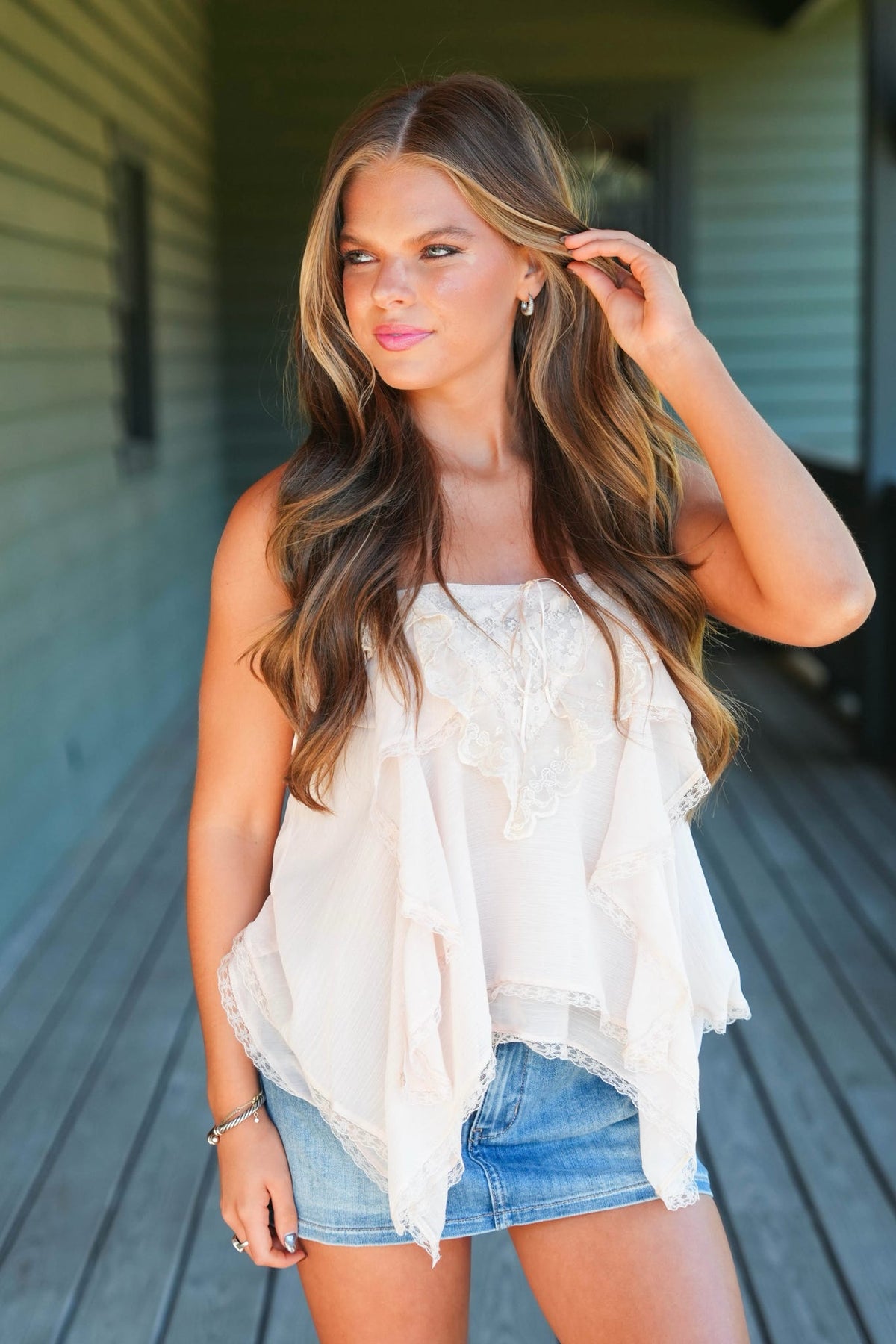 A woman modeling a white ruffled sleeveless top with denim shorts, standing on a wooden deck in front of a house.