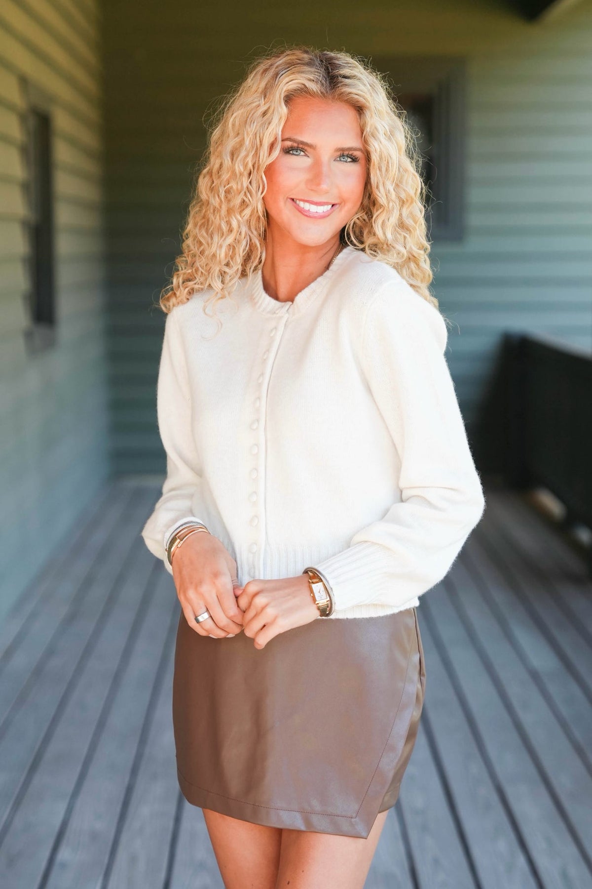 A smiling blonde woman stands on a wooden porch wearing a cream long-sleeve button-front top and a brown mini skirt.