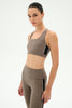 A woman modeling a matching taupe-brown activewear set consisting of a scoop-neck sports bra and high-waisted leggings, both featuring black side panels, against a plain white studio background.