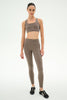 A woman modeling a matching taupe-brown activewear set consisting of a square-neck sports bra and high-waisted leggings, paired with black athletic sneakers, against a plain white studio background.
