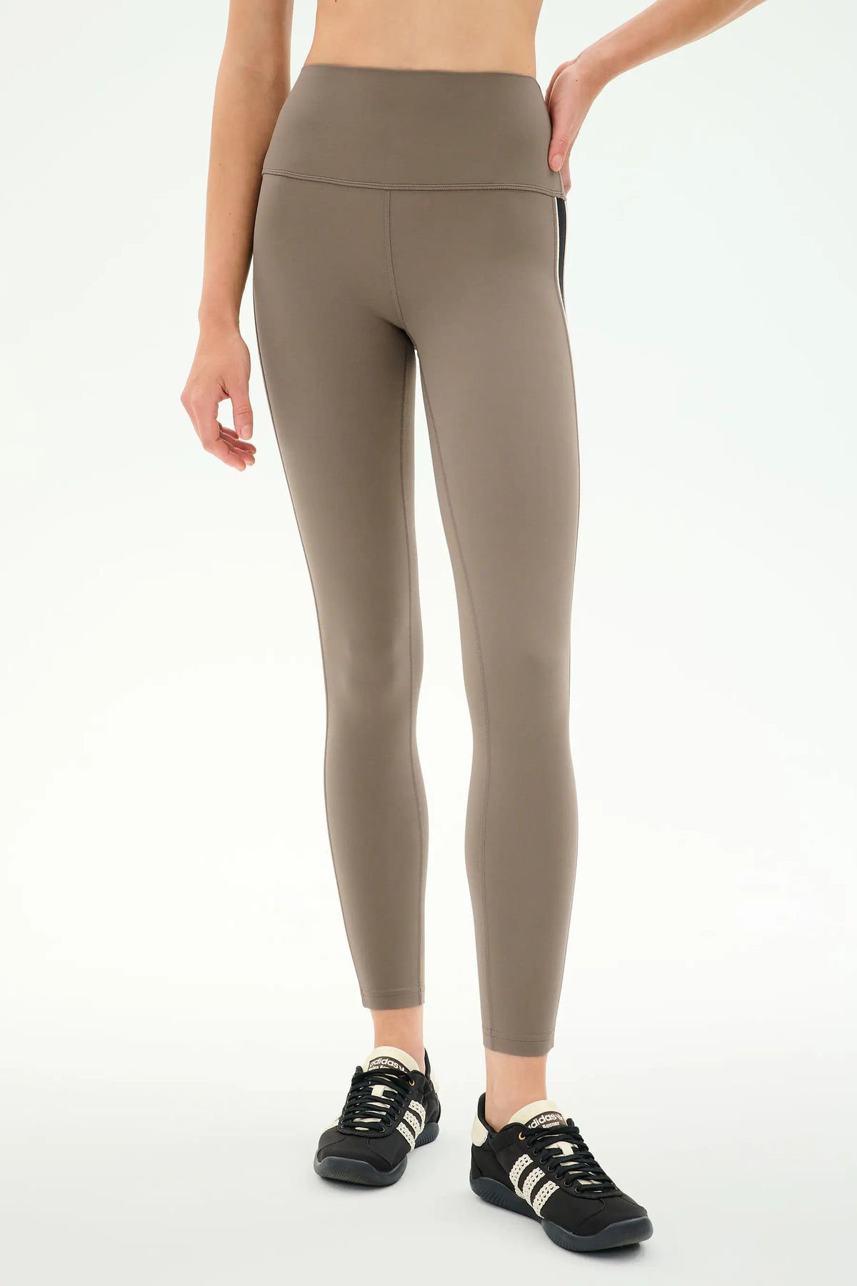 A model wearing high-waisted, full-length athletic leggings in a taupe brown color, paired with black sneakers, shown from the waist down against a plain white studio background.