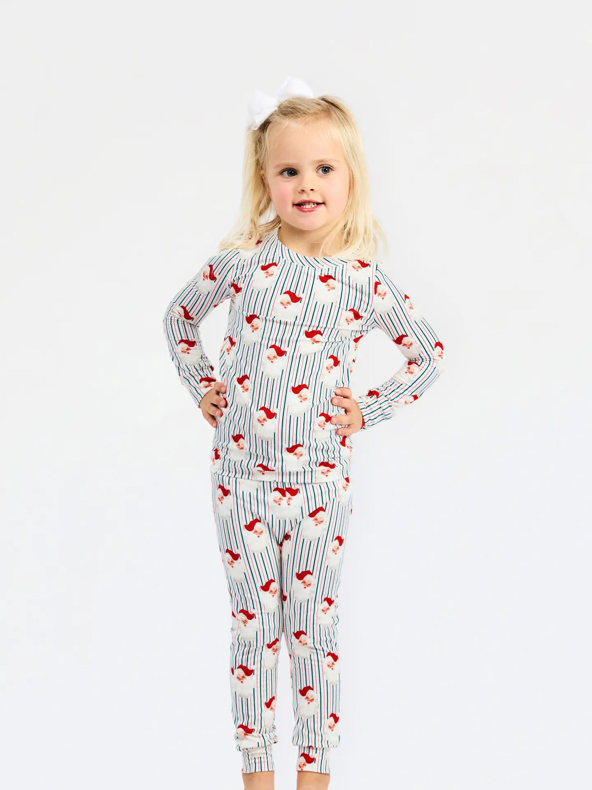 A young girl with blonde hair and a white bow stands wearing a long-sleeve pajama set with red and white pinstripes and a red Santa Claus pattern, posed against a plain white background.