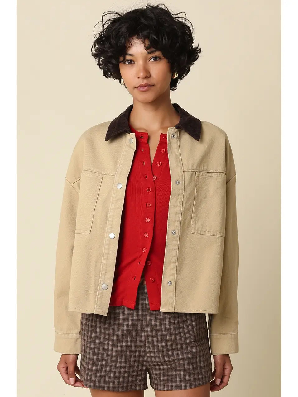 A model wearing a beige cropped utility jacket with a dark brown corduroy collar, layered over a red button-up shirt and brown plaid shorts, set against a solid beige background.