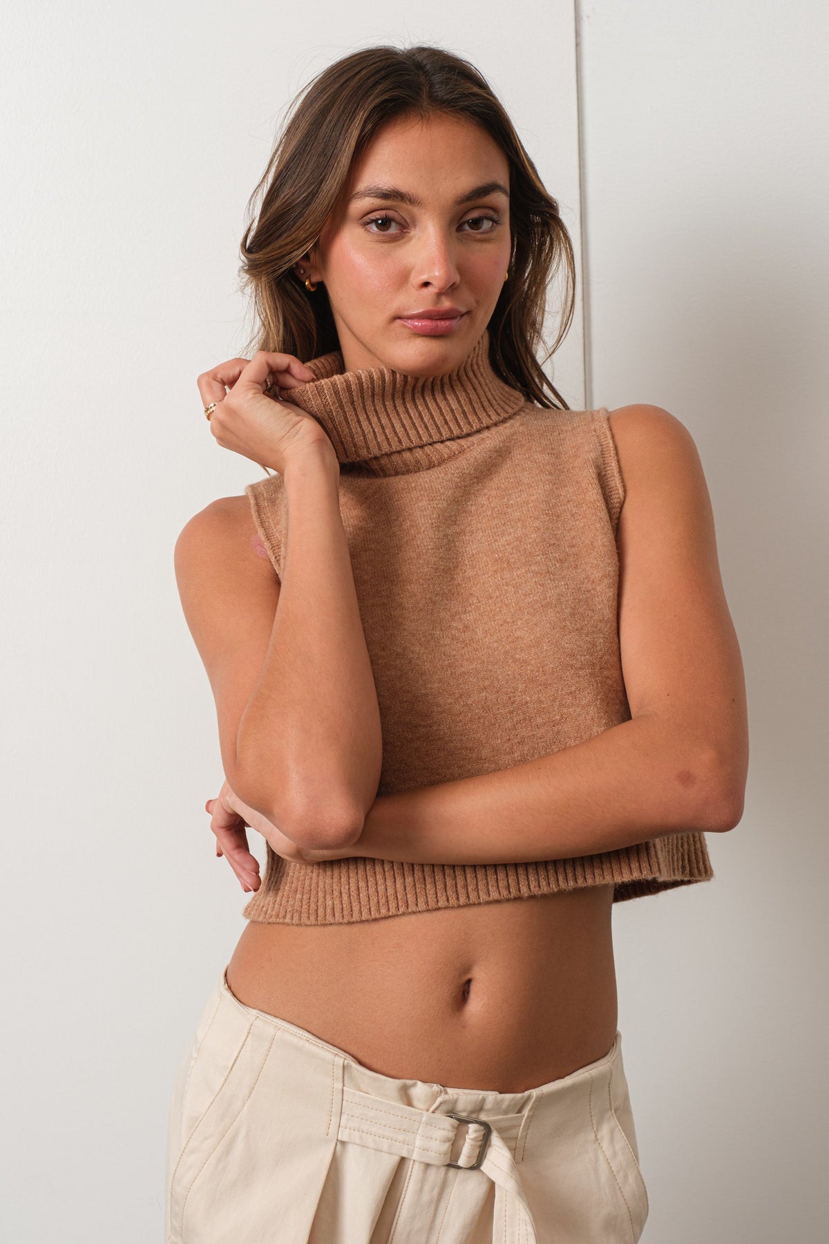 A woman wearing a sleeveless ribbed knit mock neck crop top in a warm camel brown, paired with cream-colored pants, posing against a light off-white wall.