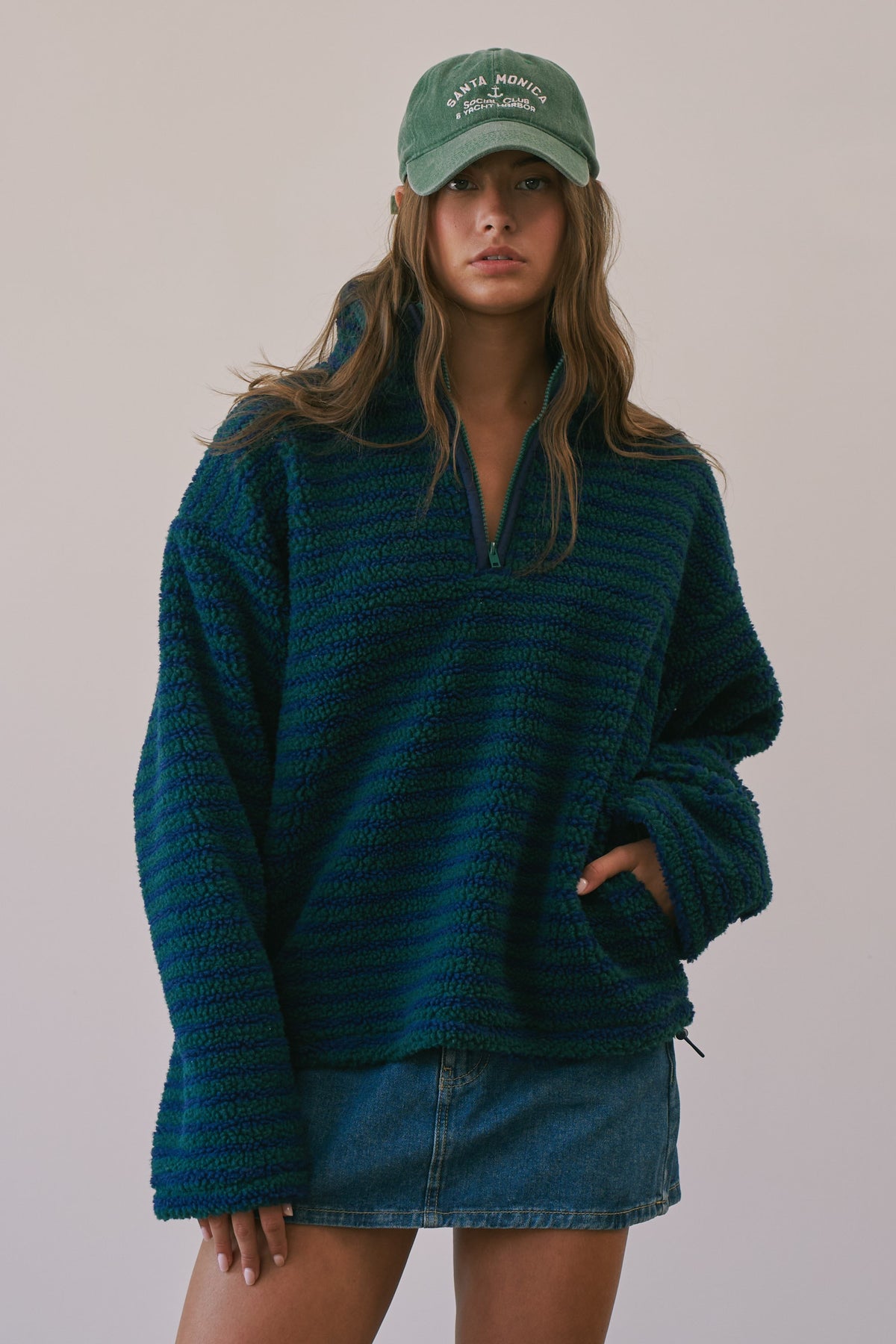 A woman wearing a dark teal textured quarter-zip fleece pullover sweater, a green baseball cap, and a denim mini skirt, standing against a plain light beige background.