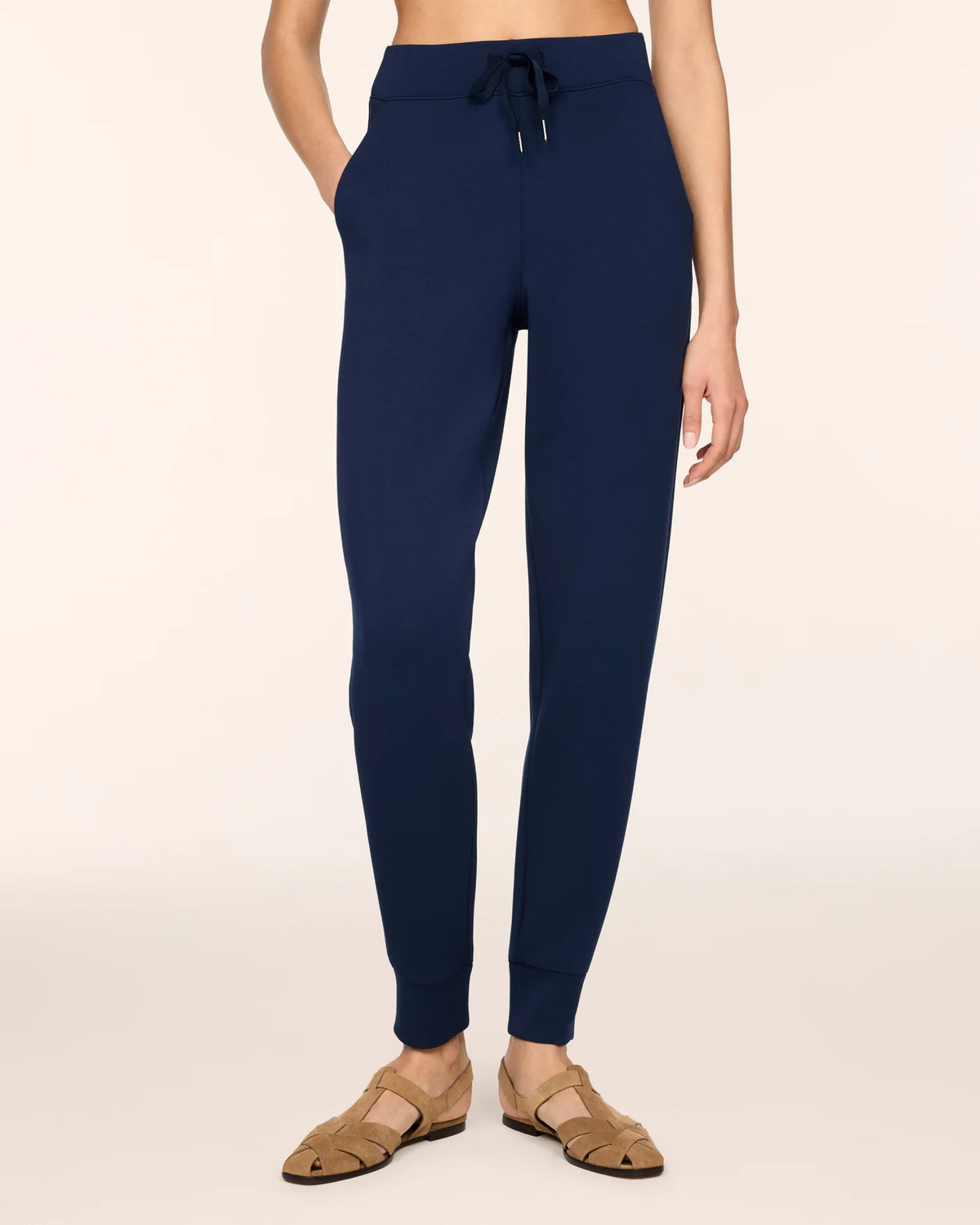A person shown wearing navy blue jogger-style pants with a drawstring waistband and ribbed ankle cuffs, paired with tan flat sandals against a plain light beige studio background.