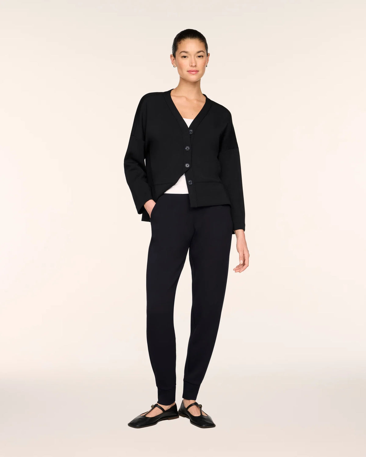 A model stands against a plain light beige studio background wearing a black cardigan with buttons over a white top, black fitted pants, and black ballet flats.