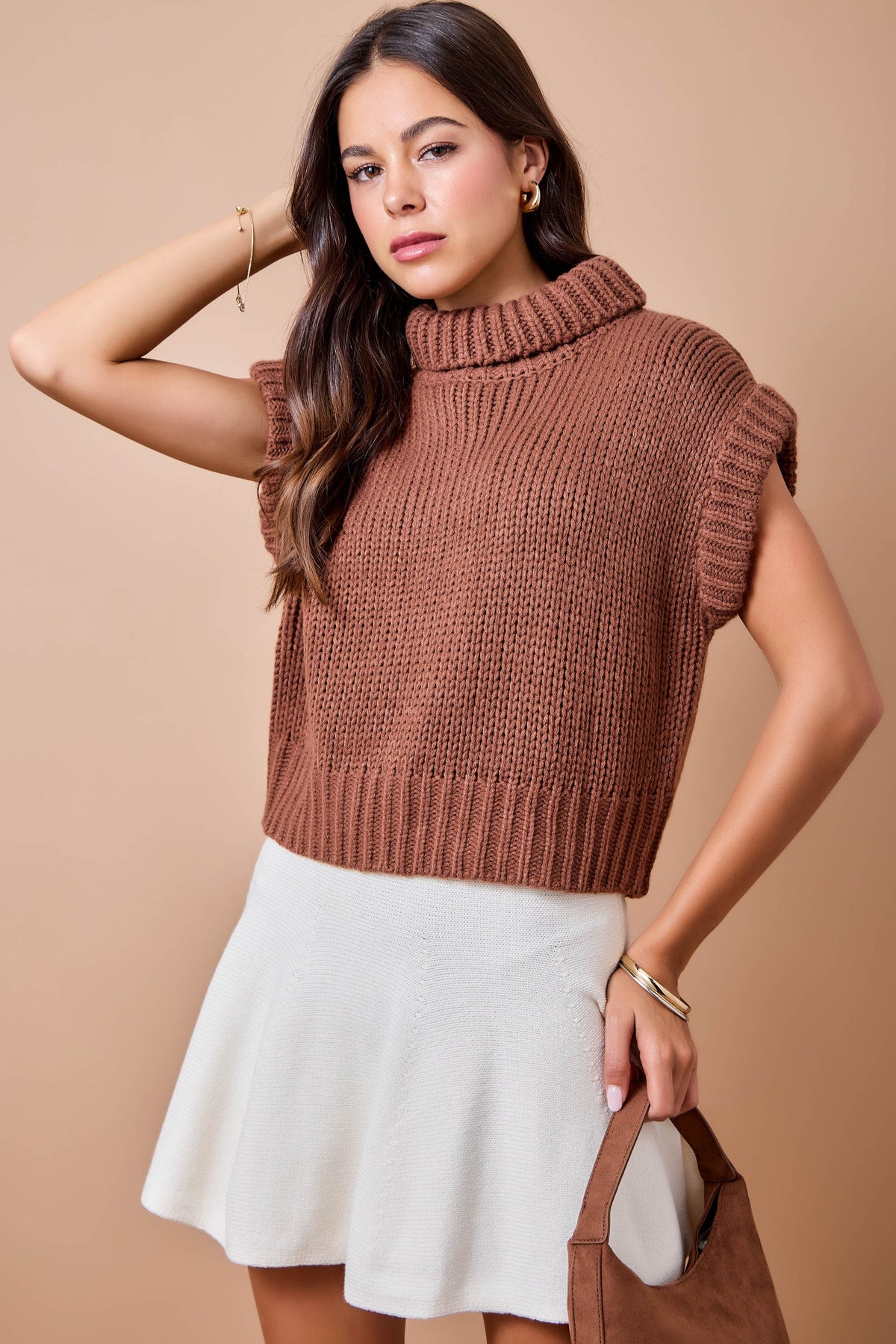 A woman models a brown ribbed knit mock neck sweater vest with short sleeves, paired with a white flared skirt and holding a brown leather handbag against a solid beige background.