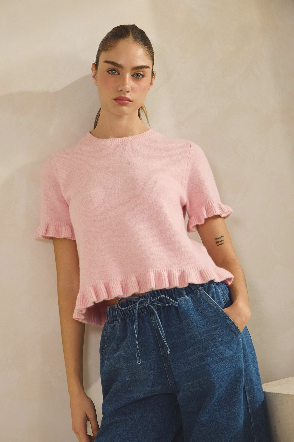 Woman wearing a pink ruffled top and blue pants against a neutral background