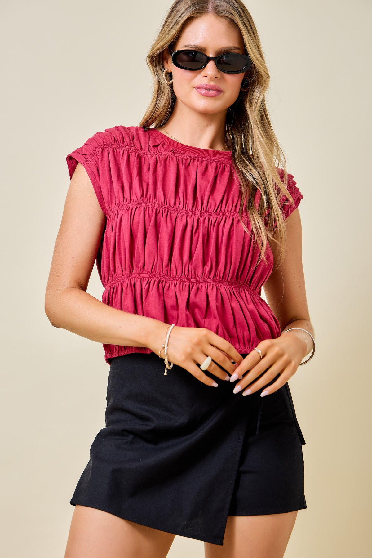 A woman modeling a sleeveless, smocked-embellished magenta top paired with black wrap-style shorts against a solid beige background.