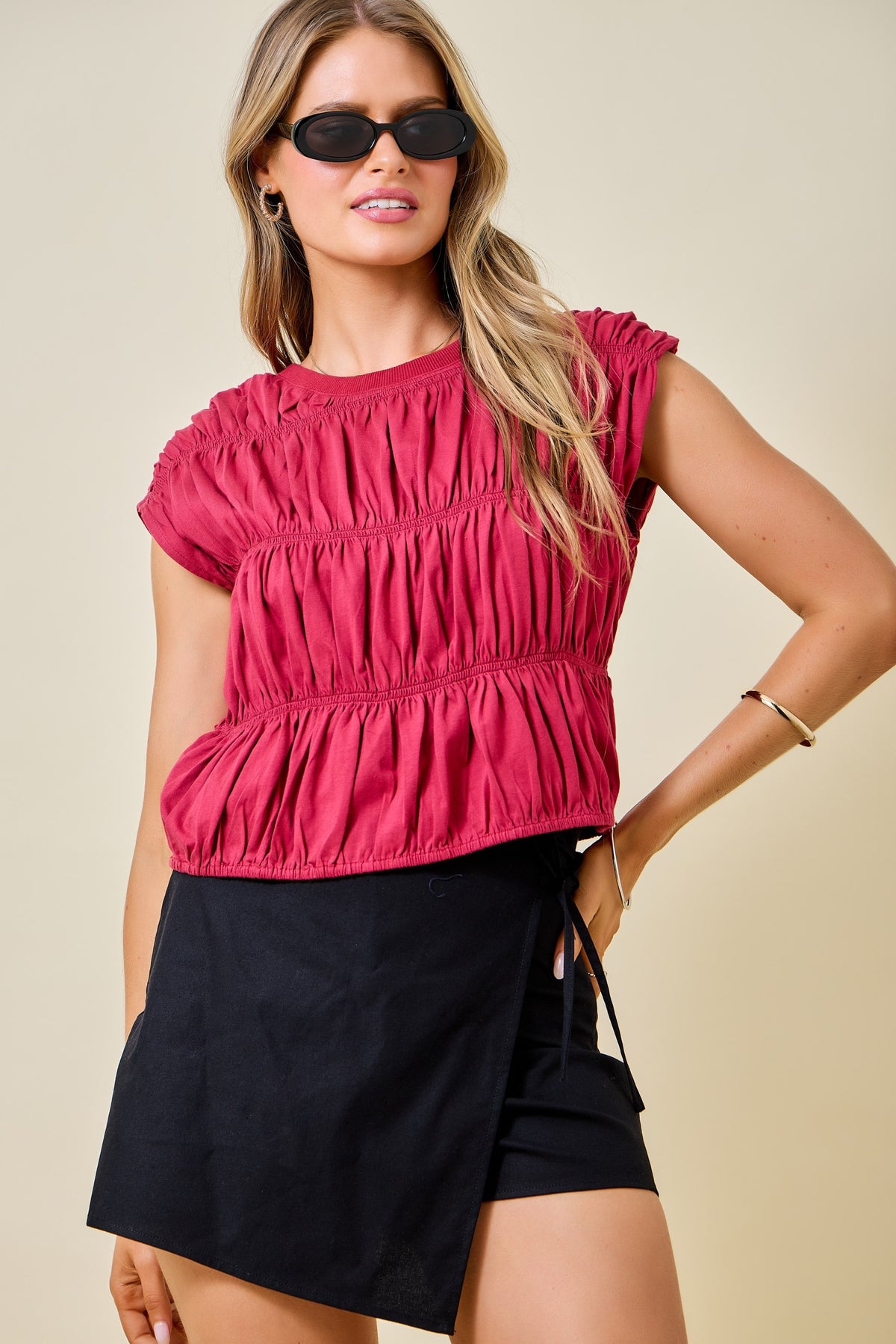 A woman modeling a bright pinkish-red cropped short-sleeve top with horizontal ruching, paired with black wrap-style shorts, against a plain beige studio background.