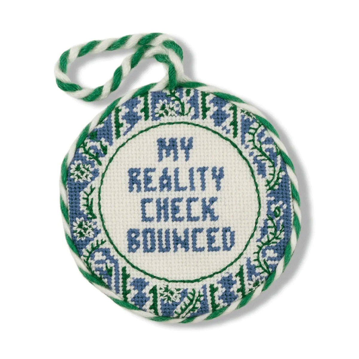 A round cross-stitch ornament featuring the text 'MY REALITY CHECK BOUNCED' in blue thread, surrounded by a blue and green floral border, with a green and white striped hanging loop at the top, shown on a plain white background.