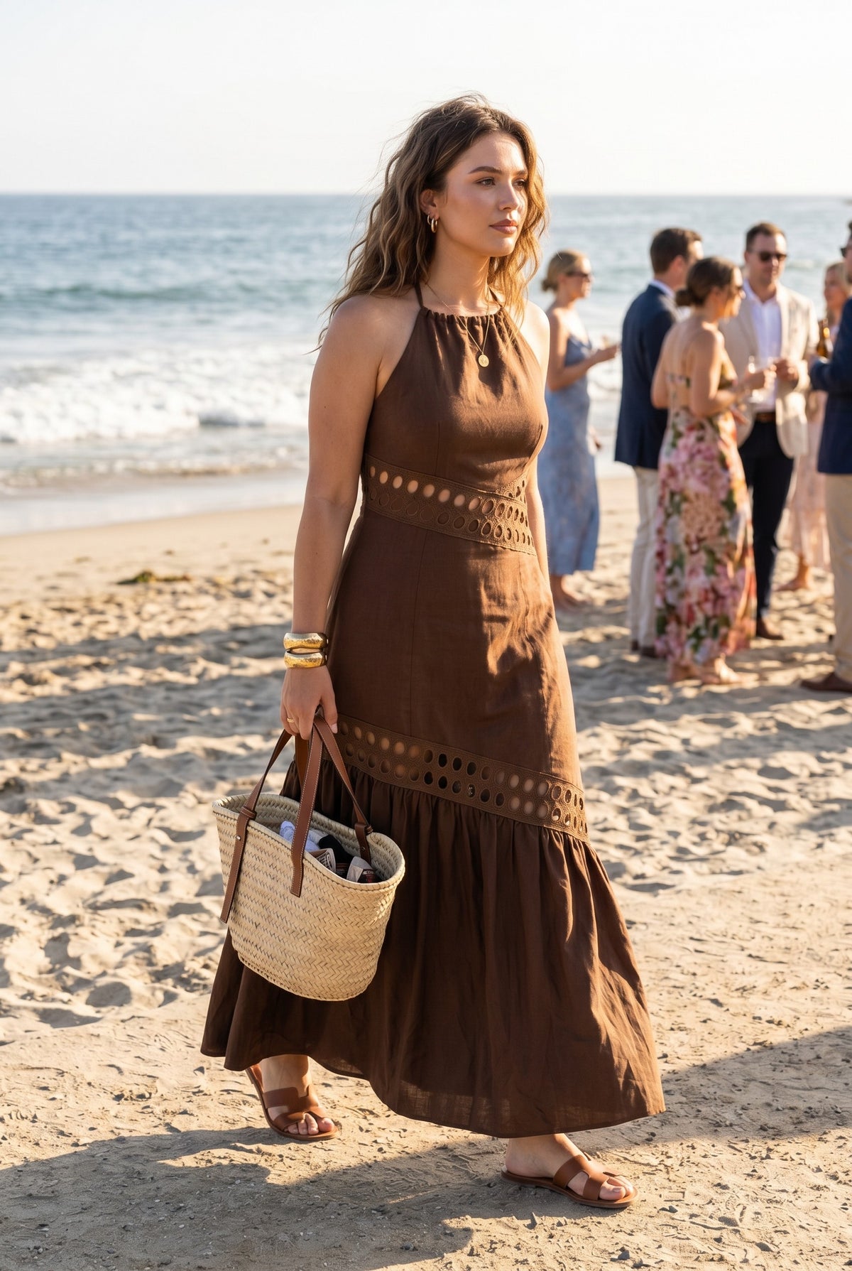 Woman in a brown dress holding a straw bag on a beach with ocean and people in the background