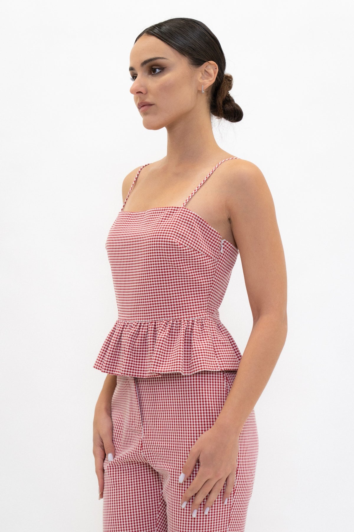 A woman modeling a matching pink and white gingham two-piece set, featuring a sleeveless camisole top with a peplum waist and coordinating pants, shot against a plain white studio background.