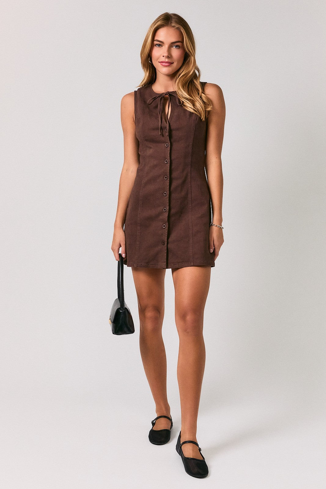 A woman modeling a sleeveless brown mini dress with a front button placket and tie neckline, paired with black flats and holding a small black handbag, against a light gray studio background.