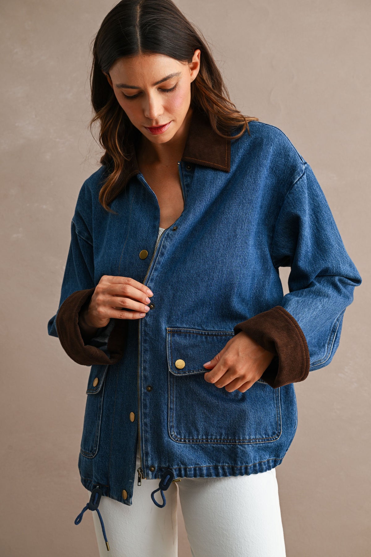 A woman wearing an oversized blue denim jacket with contrasting brown suede-like collar and cuff trim, paired with white pants, set against a neutral beige background.