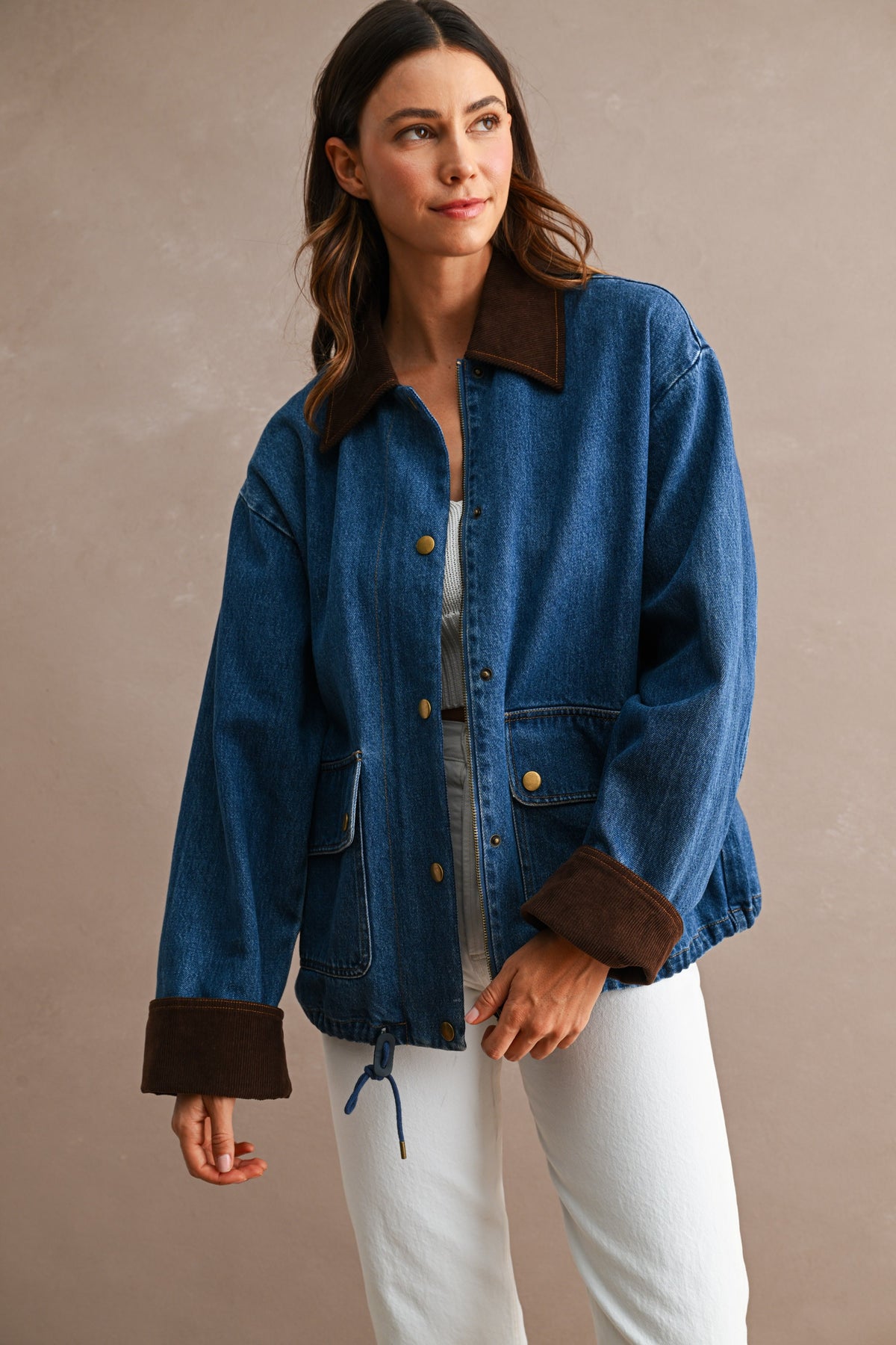 A woman wearing a blue denim jacket with dark brown suede-like collar, cuffs, and front pockets, paired with white pants, standing against a textured beige studio background.