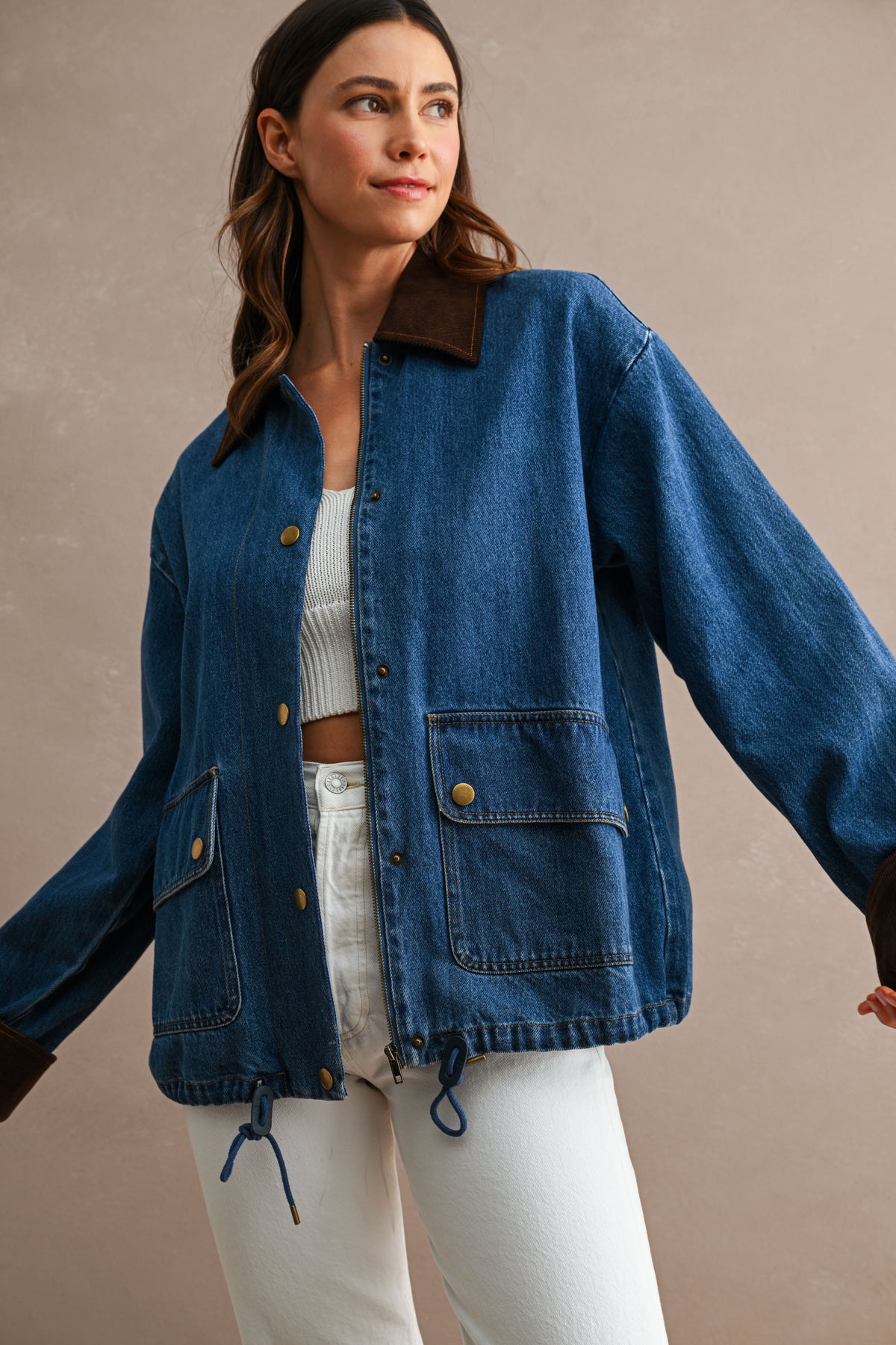 A woman wearing a blue denim jacket with dark brown corduroy collar and cuffs, paired with white pants and a white ribbed crop top, standing against a neutral beige background.