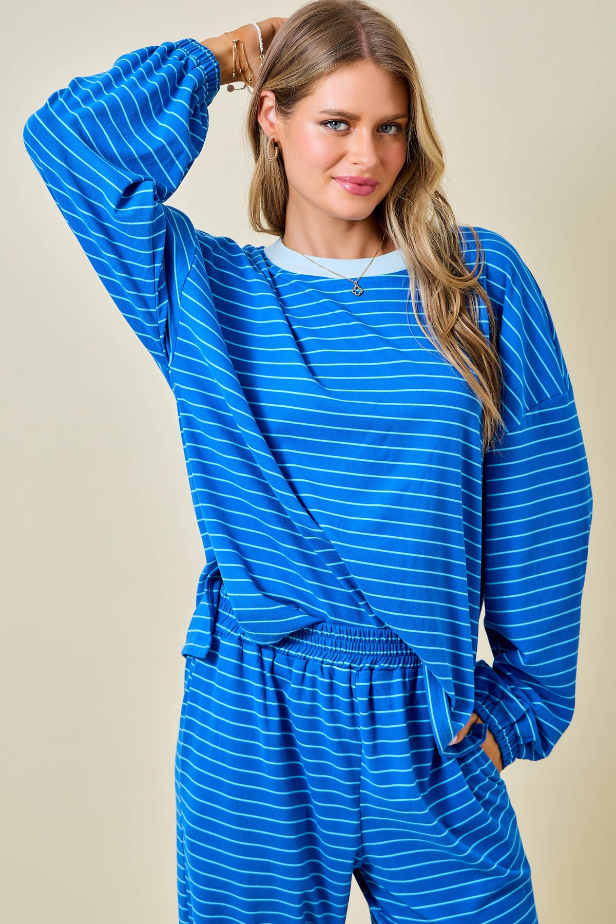 A woman modeling a matching blue and white striped lounge set, featuring a long-sleeve top and pants, against a solid beige background.