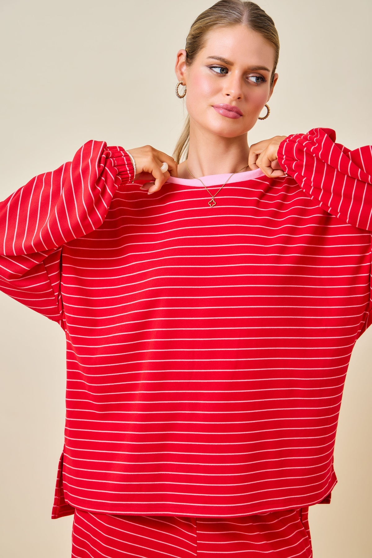 A woman wearing a red long-sleeve top with thin white horizontal stripes, holding the collar open to show a light pink inner layer. She is also wearing matching red and white striped pants against a solid beige background.