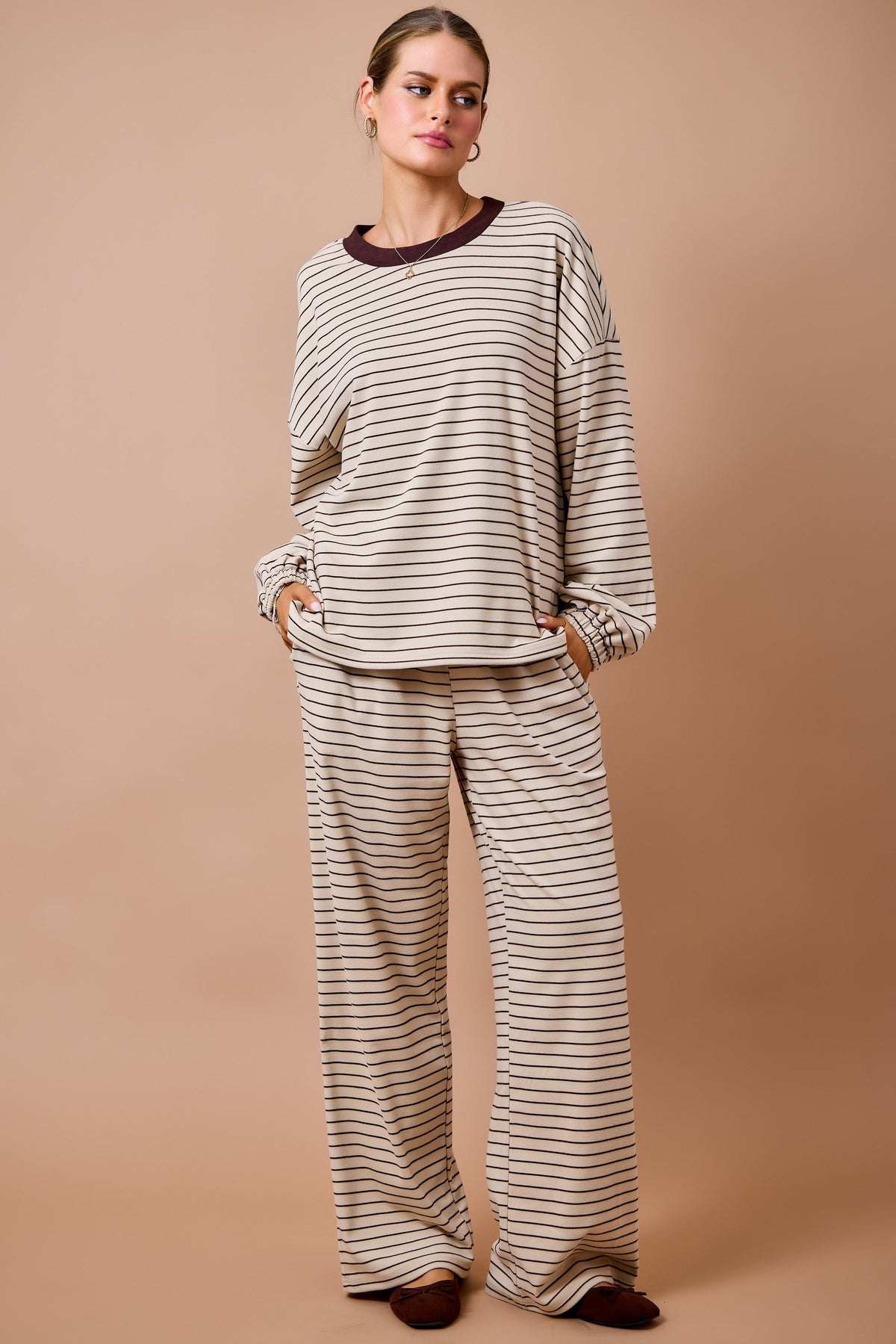 A woman modeling a matching two-piece loungewear set consisting of a long-sleeve top and wide-leg pants, both in a beige and dark brown horizontal stripe pattern, against a solid tan studio background.