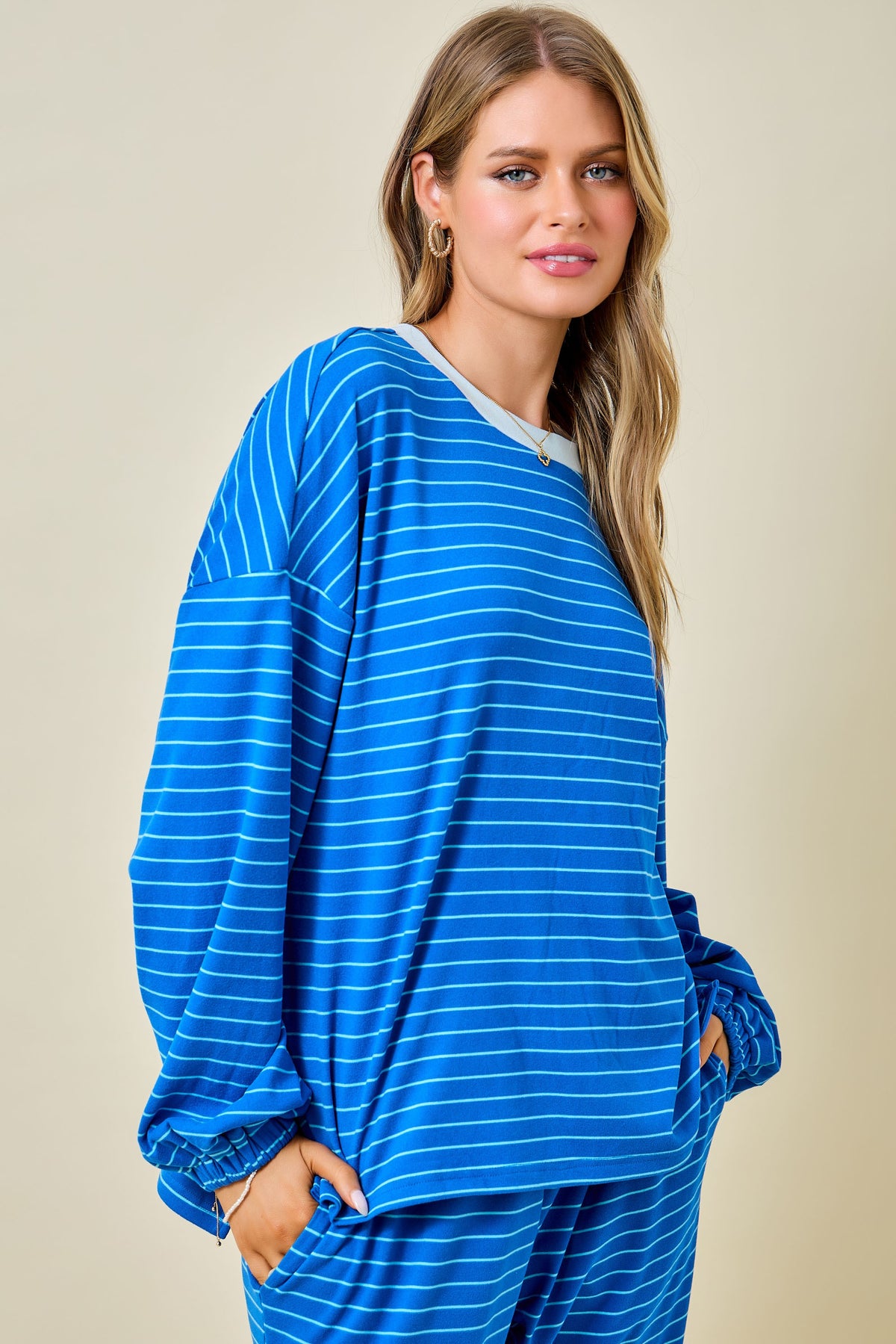 A woman modeling a matching blue loungewear set with thin white horizontal stripes, featuring a long-sleeve top and pants, against a solid beige background.