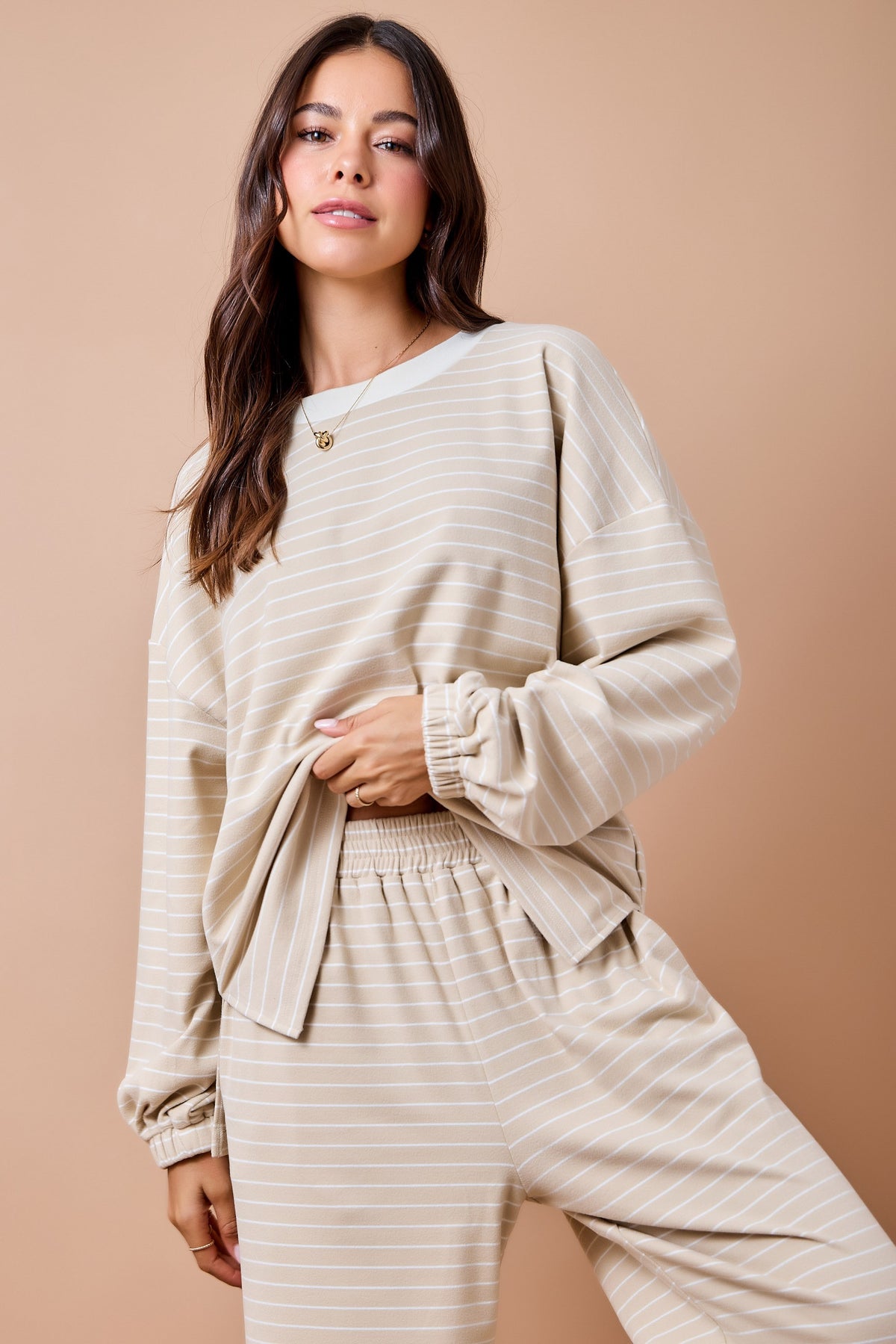 A woman modeling a matching beige and white striped lounge set, featuring a long-sleeve top with gathered cuffs and matching wide-leg pants, set against a solid warm beige background.