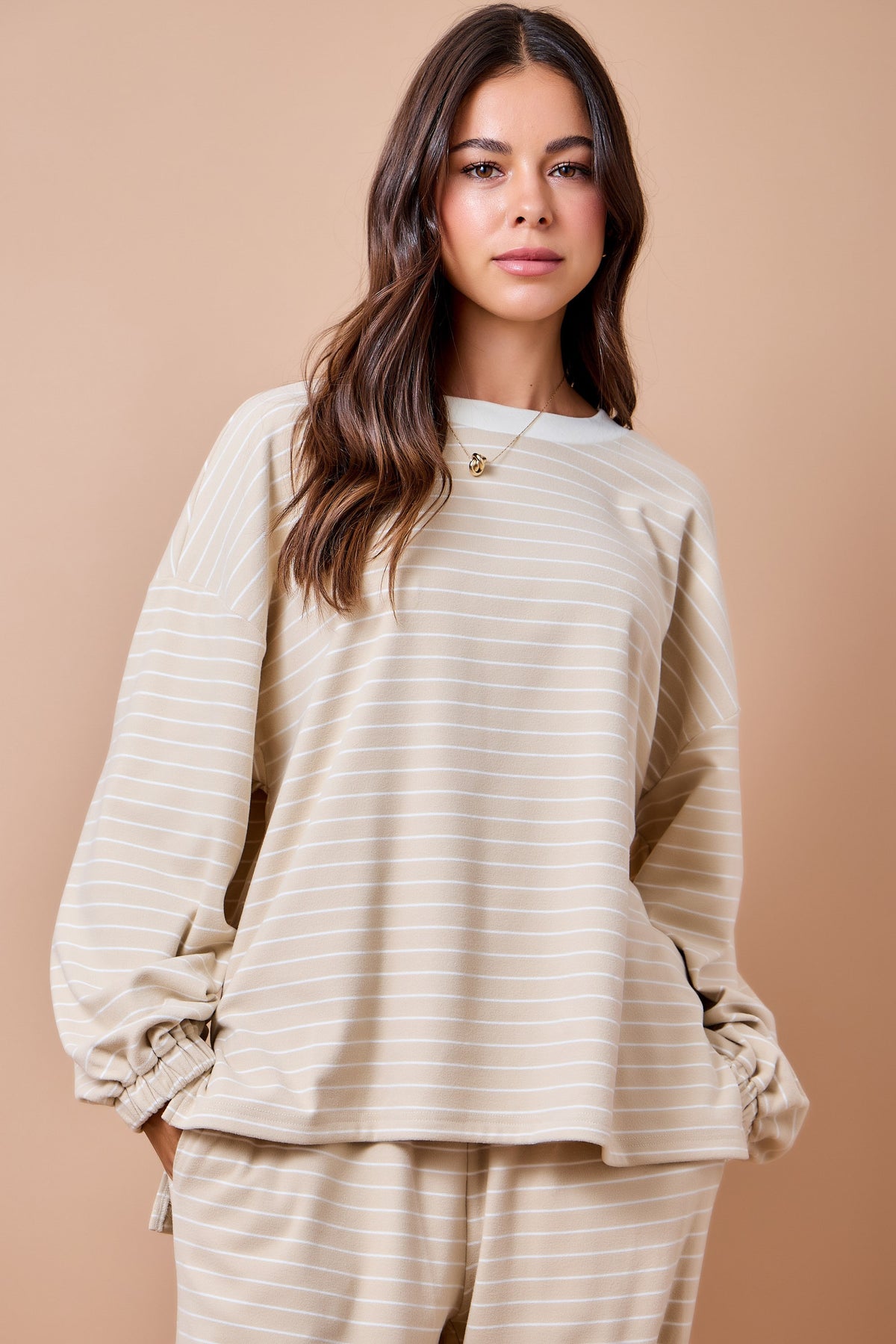 A woman modeling a beige and white striped lounge set, featuring a long-sleeve top with gathered cuffs and matching pants, set against a solid beige background.