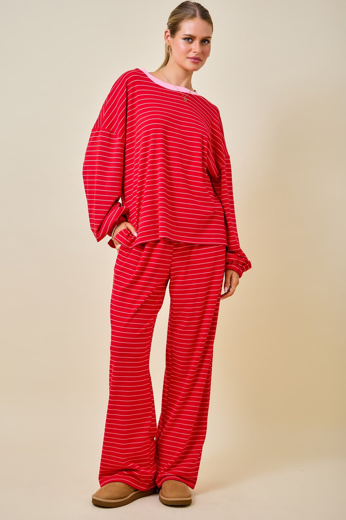 A woman modeling a red and white striped two-piece lounge set consisting of a long-sleeve top and matching wide-leg pants, paired with brown slippers against a solid beige studio background.