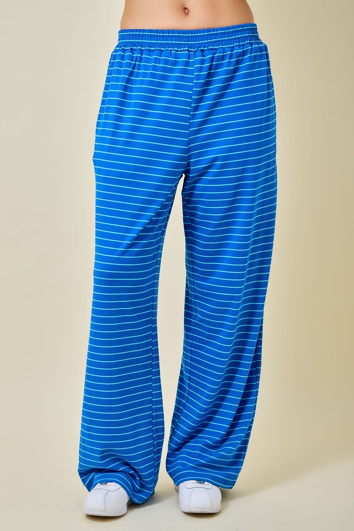 Blue wide-leg sweatpants with thin white horizontal stripes, shown on a model from the waist down against a solid beige background.