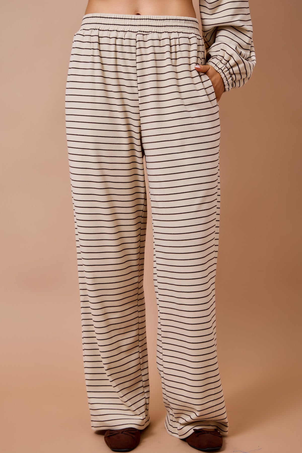 A person wearing loose, wide-leg pants with thin horizontal black stripes on a warm cream background, paired with a matching striped long-sleeve top. The photo is cropped from the lower torso to the feet against a plain beige studio backdrop.