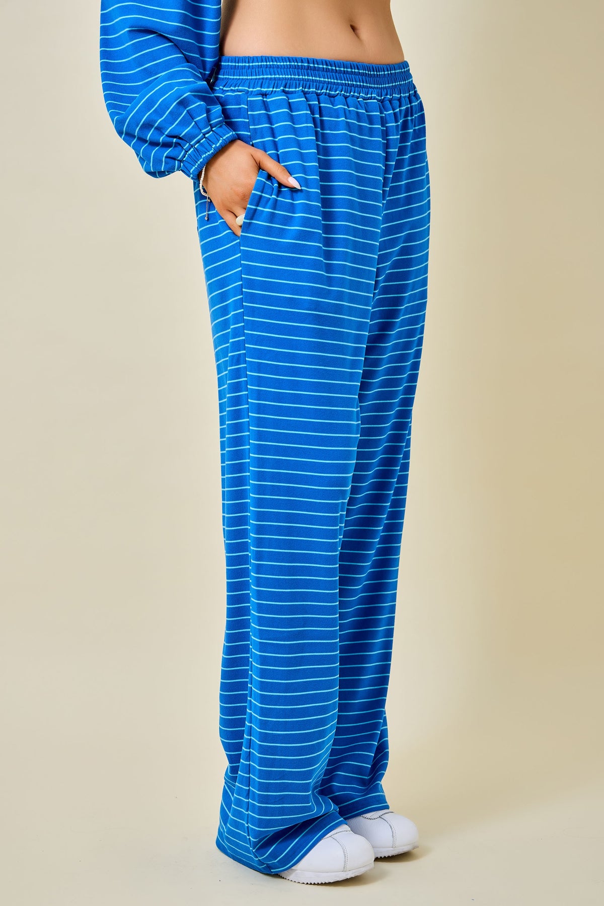 A person wearing loose blue pants with thin white horizontal stripes, shown from the waist down against a plain beige studio background.