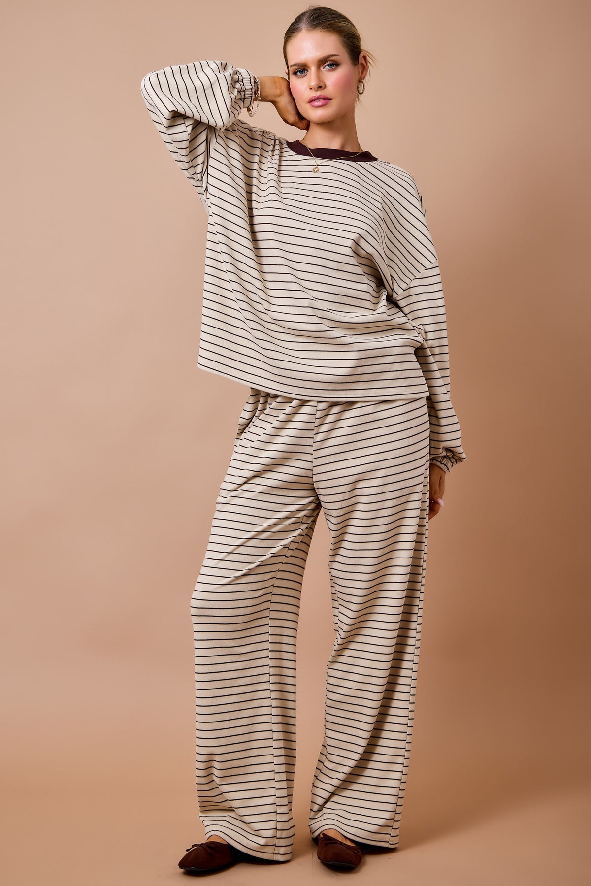 A woman modeling a matching two-piece loungewear set featuring a beige and brown striped long-sleeve top and wide-leg pants, posed against a solid beige studio background.