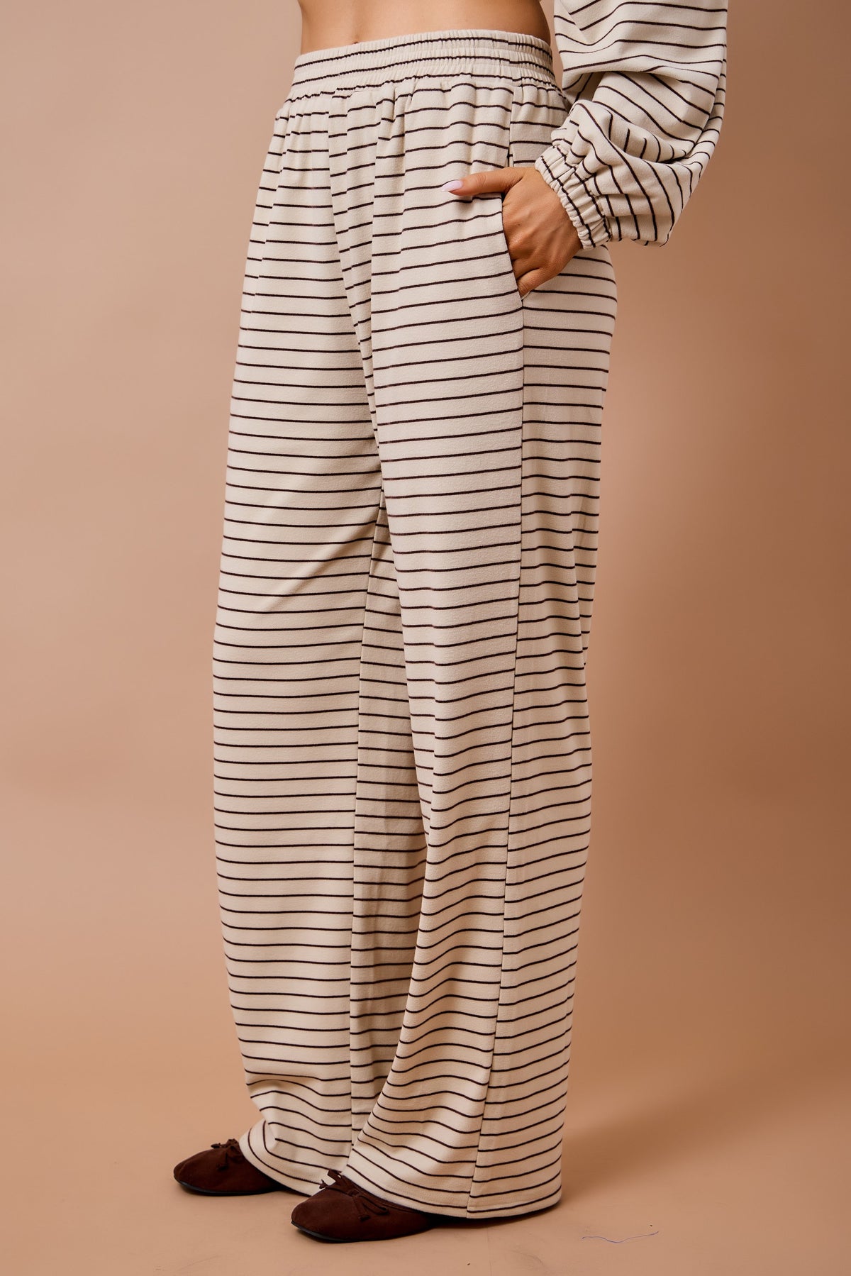 A person wearing loose, wide-leg pants with thin horizontal black stripes on a warm beige base, shown from the waist down against a solid tan studio background.