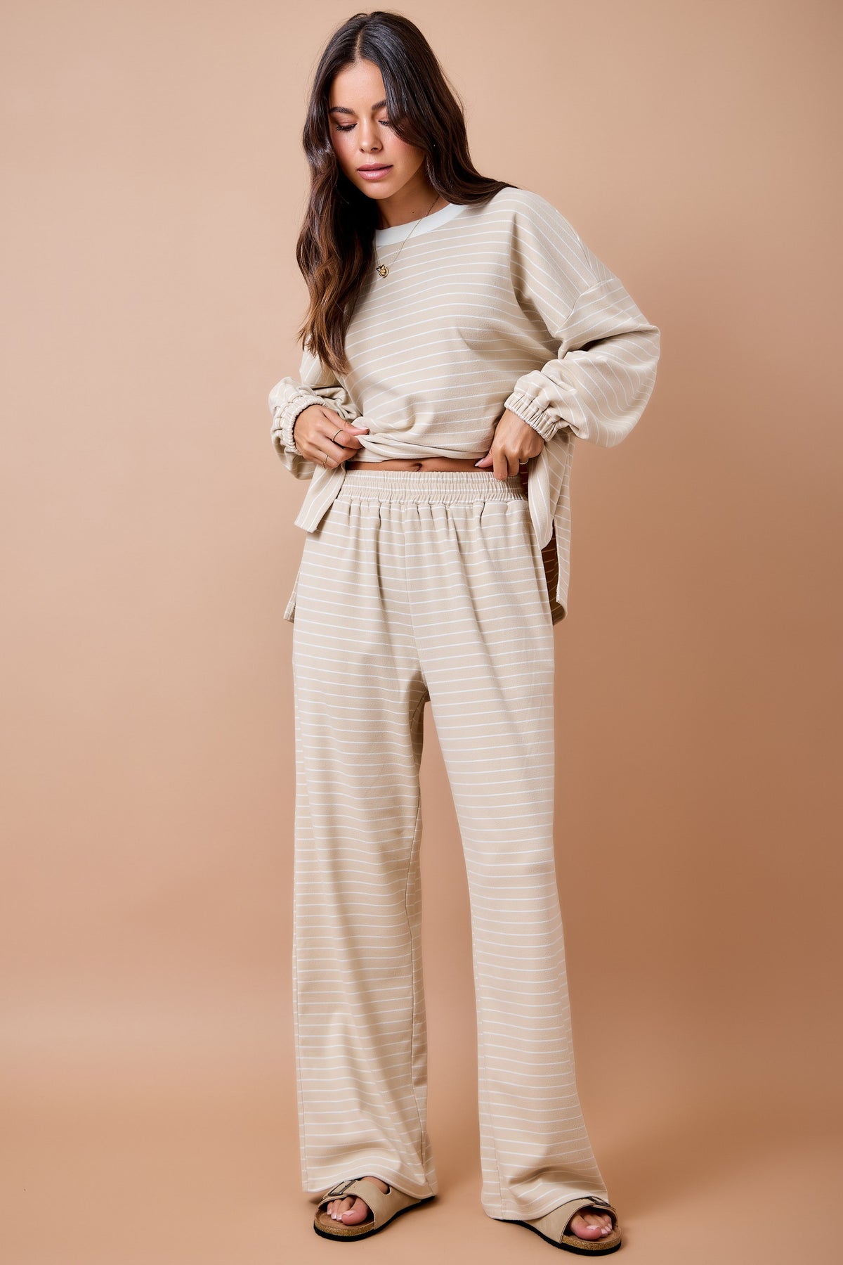 A woman modeling a matching beige and white striped lounge set, featuring a long-sleeve cropped top and wide-leg pants, against a solid warm beige studio background.