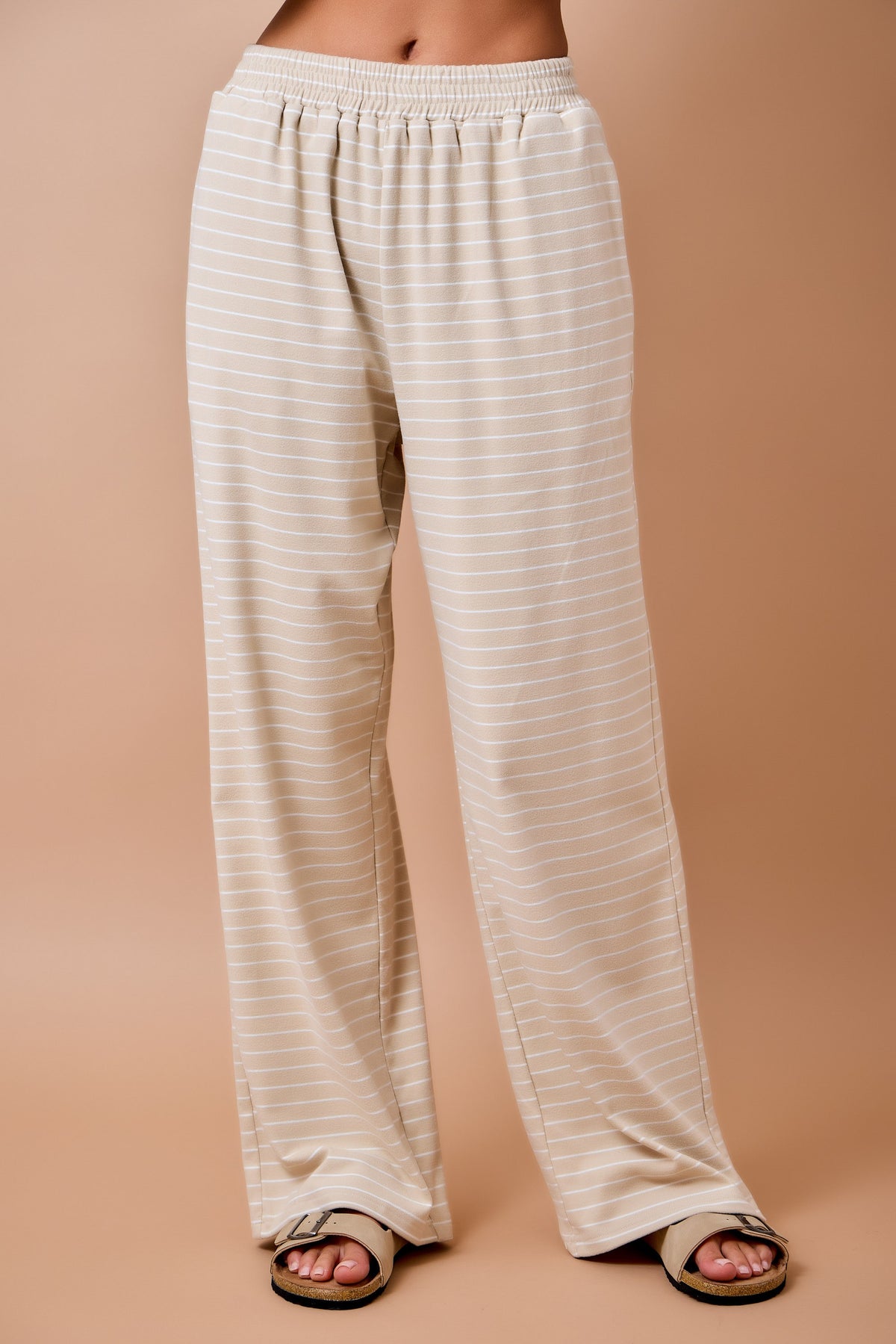 A person wearing loose, wide-leg pants with thin horizontal beige and off-white stripes, shown from the waist down against a solid warm tan studio background.