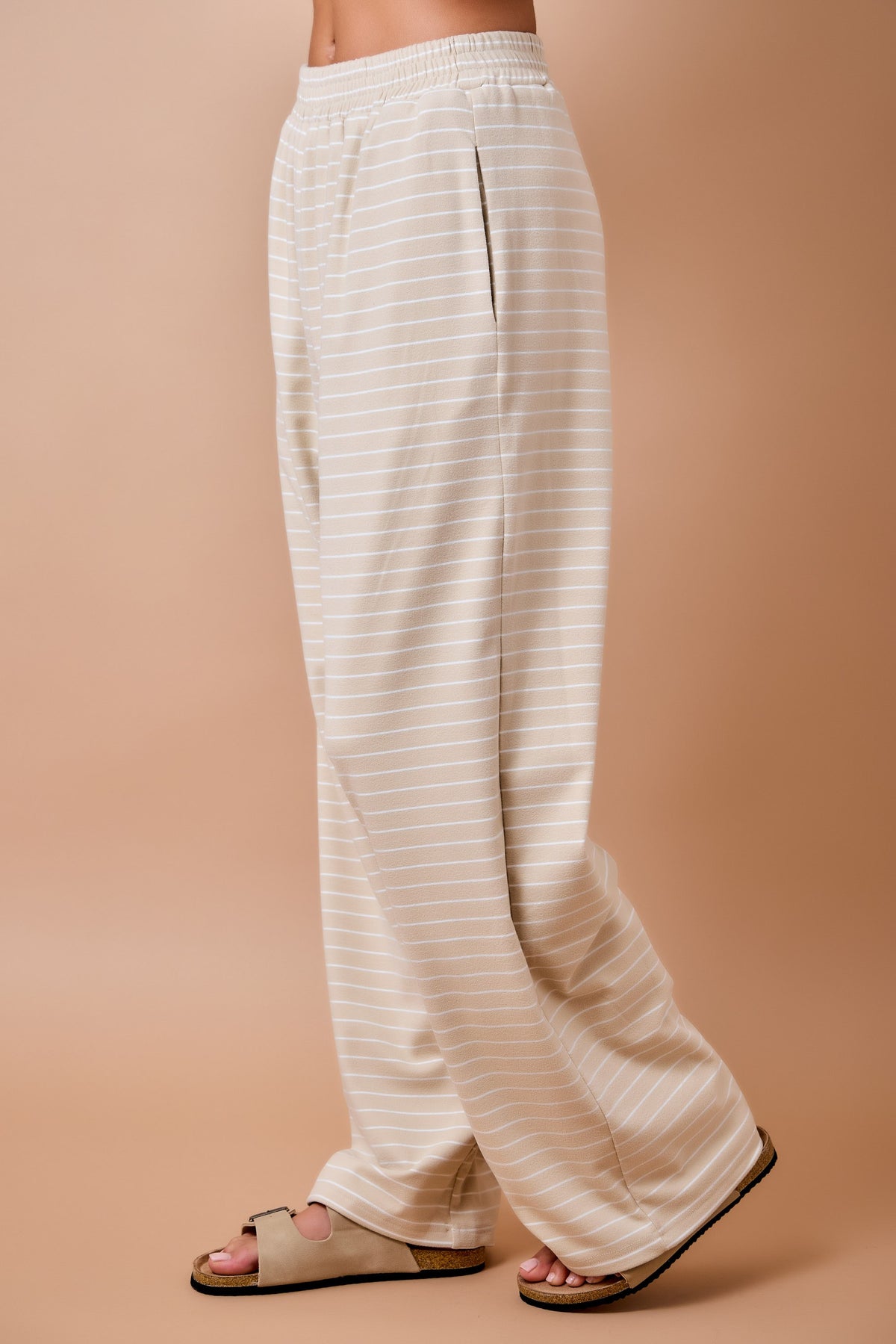 A person wearing loose, wide-leg pants with thin horizontal stripes in cream and beige, paired with tan slide sandals, against a solid warm beige studio background.