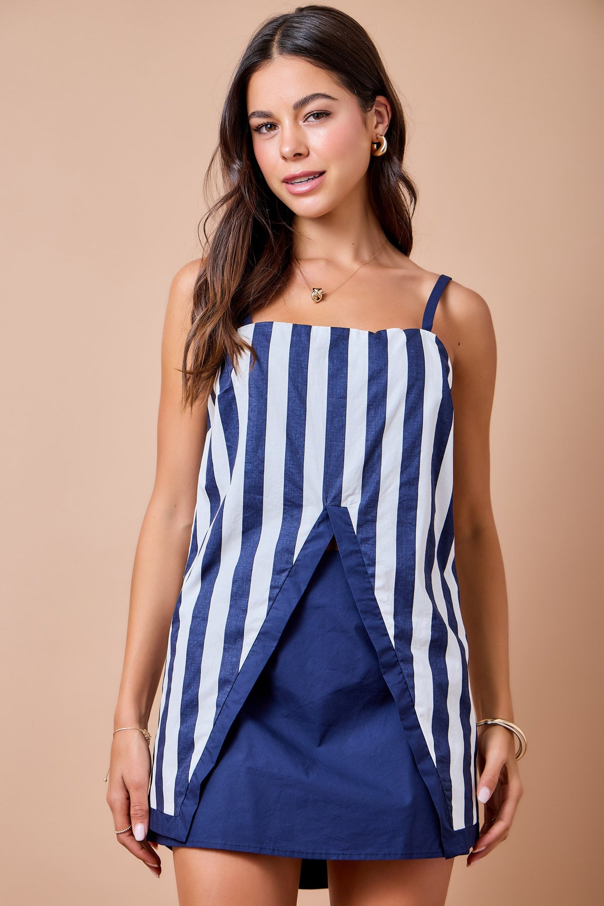 A woman modeling a sleeveless navy and white vertically striped mini dress with a wrap-style front overlay, set against a solid beige studio background.