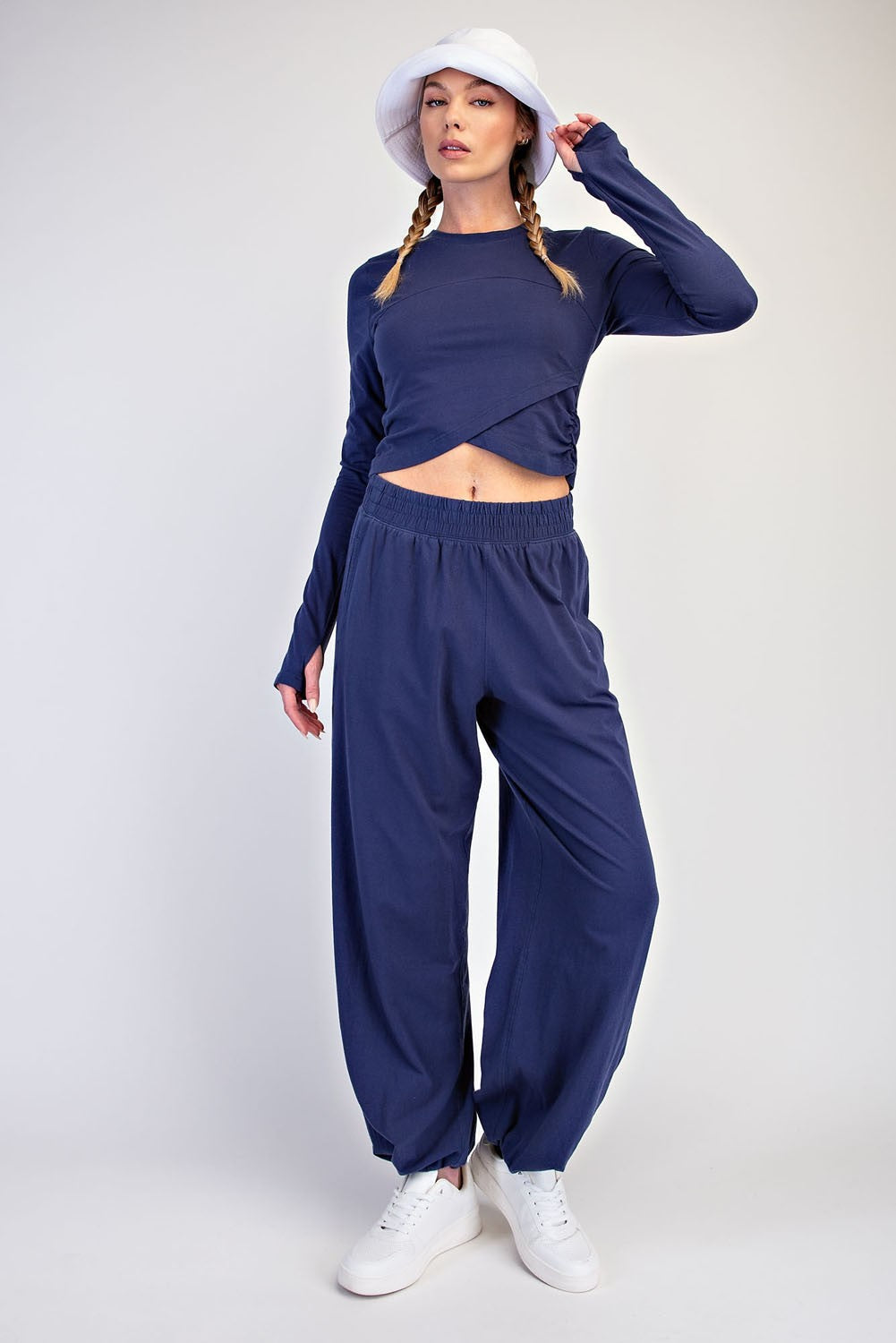 A model wearing a navy blue long-sleeve crop top and matching wide-leg pants, styled with a white bucket hat and white sneakers against a light gray studio background.
