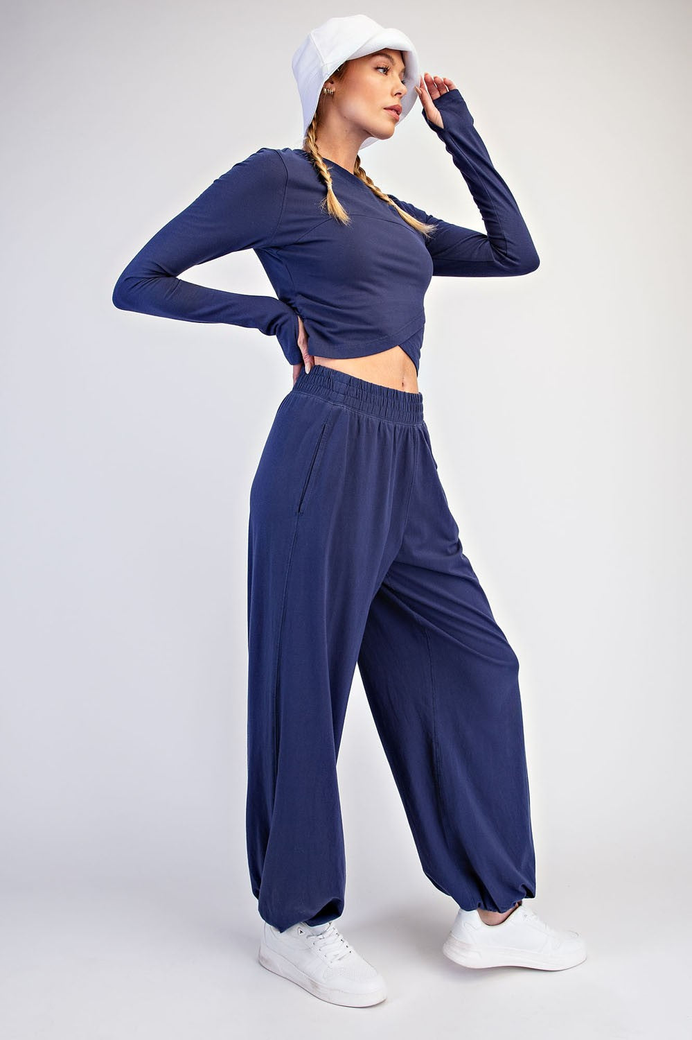 A woman modeling a navy blue two-piece activewear set consisting of a long-sleeve crop top and matching wide-leg jogger pants, styled with a white bucket hat and white sneakers against a light gray studio background.