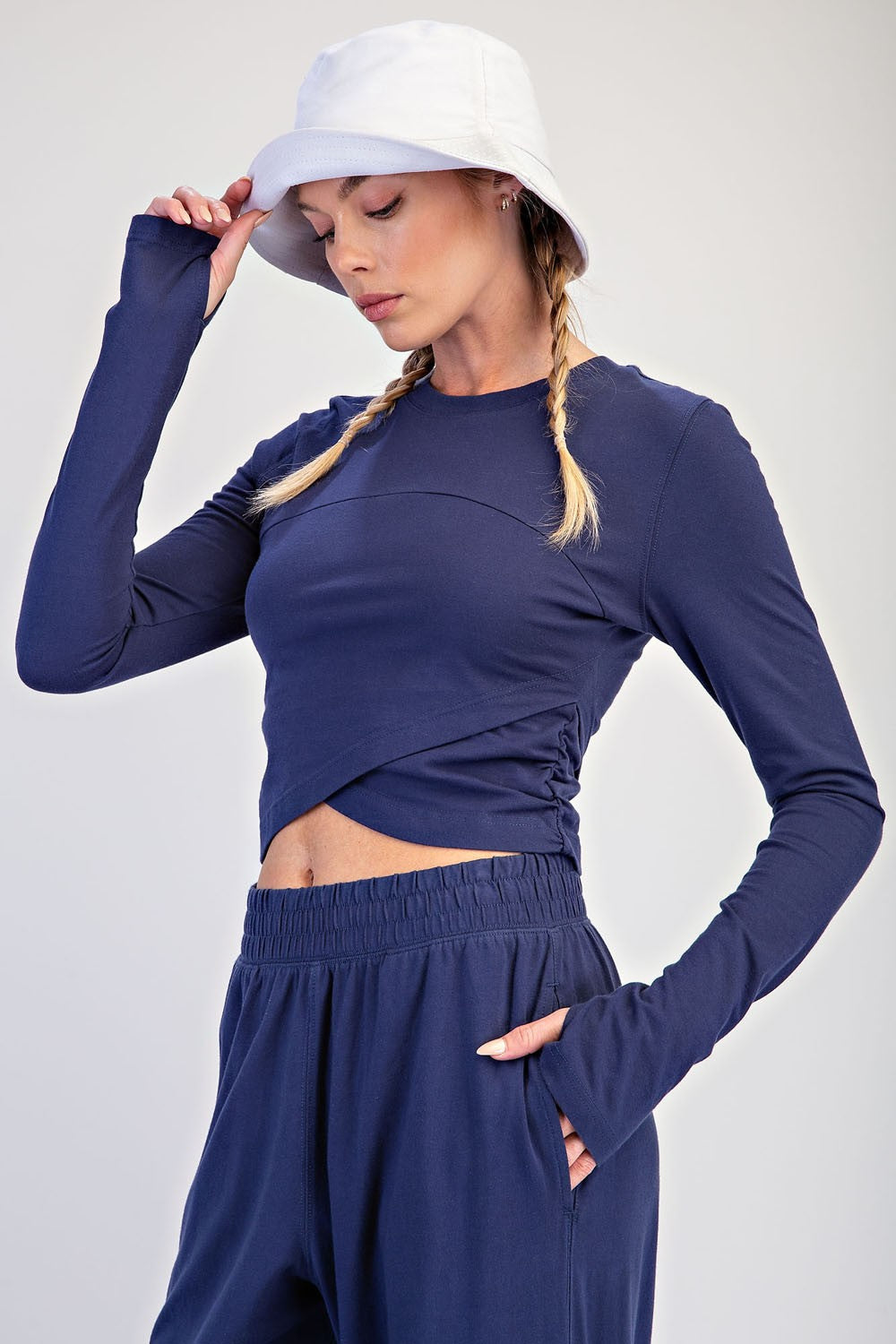 A woman wearing a navy blue long-sleeve crop top and matching high-waisted pants, styled with a white bucket hat, against a light gray studio background.